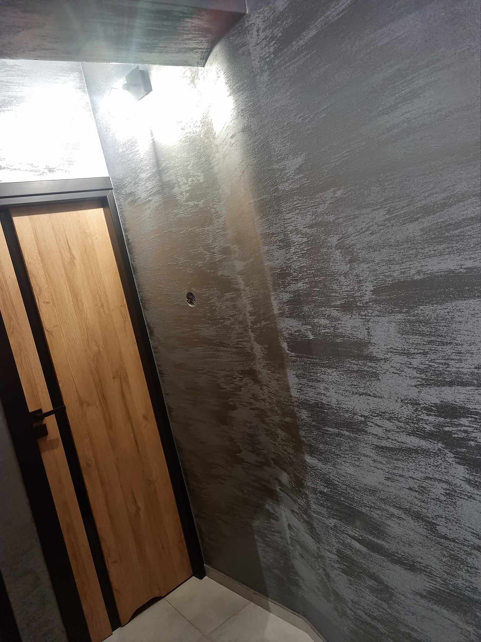 Ściana w szarym odcieniu ze strukturą imitującą przecierkę, obok drewniane drzwi z czarną ramą i klamką, widoczny fragment sufitu z lampą.