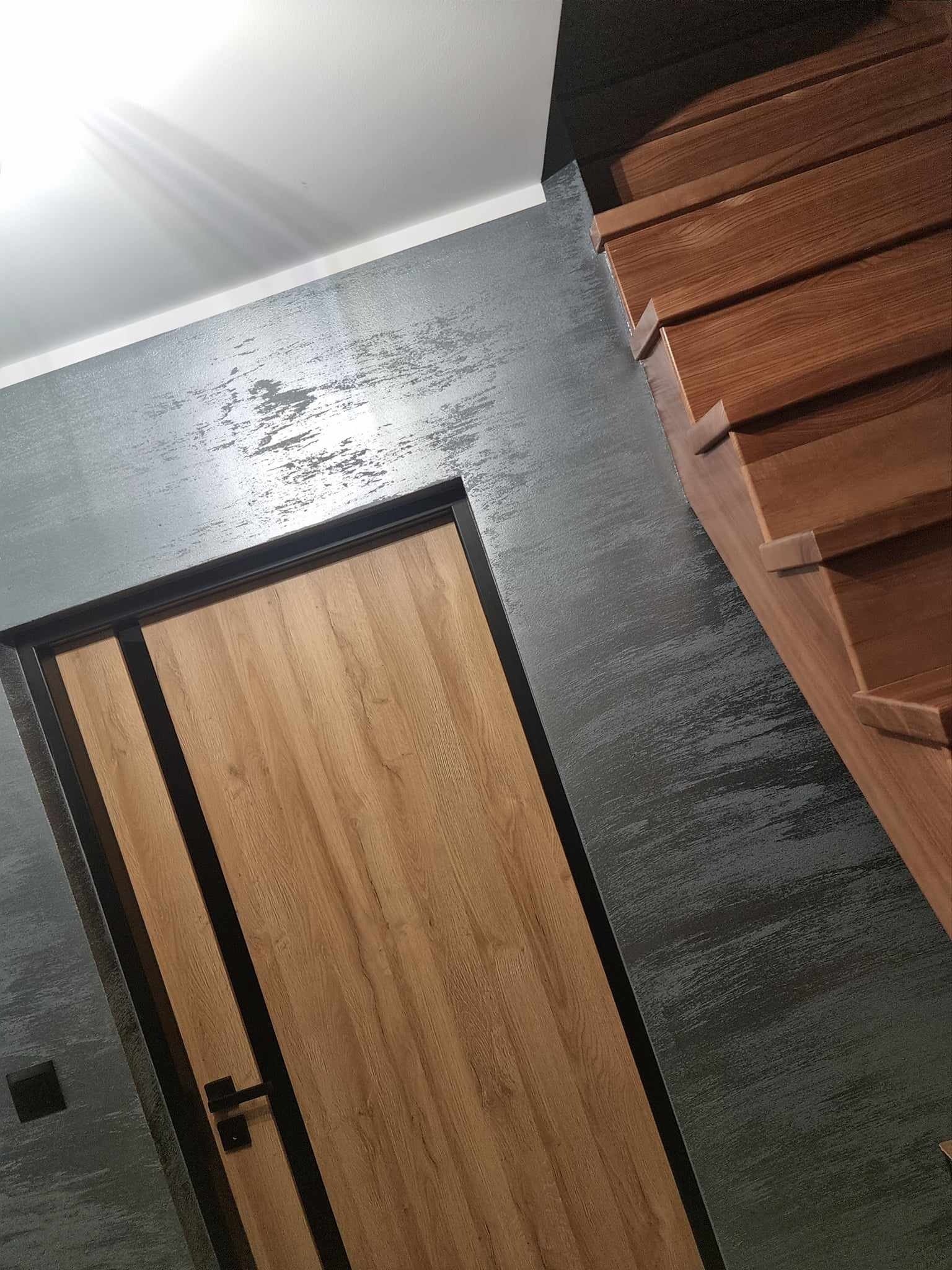 Wnętrze z drewnianymi schodami, drzwiami z okleiną w kolorze dębu i ścianą pokrytą dekoracyjnym tynkiem w odcieniach szarości.