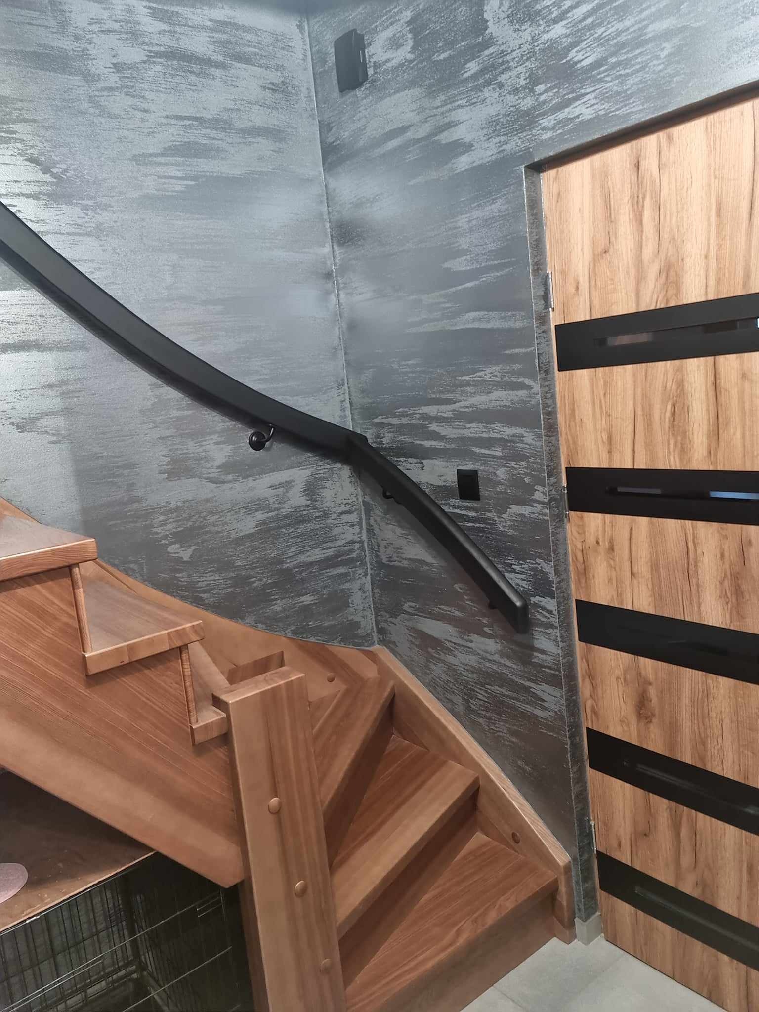 Drewniane schody z czarną, zakrzywioną poręczą na tle ściany z efektem dekoracyjnym w odcieniach szarości, obok drzwi z okleiną drewnopodobną z czarnymi akcentami.