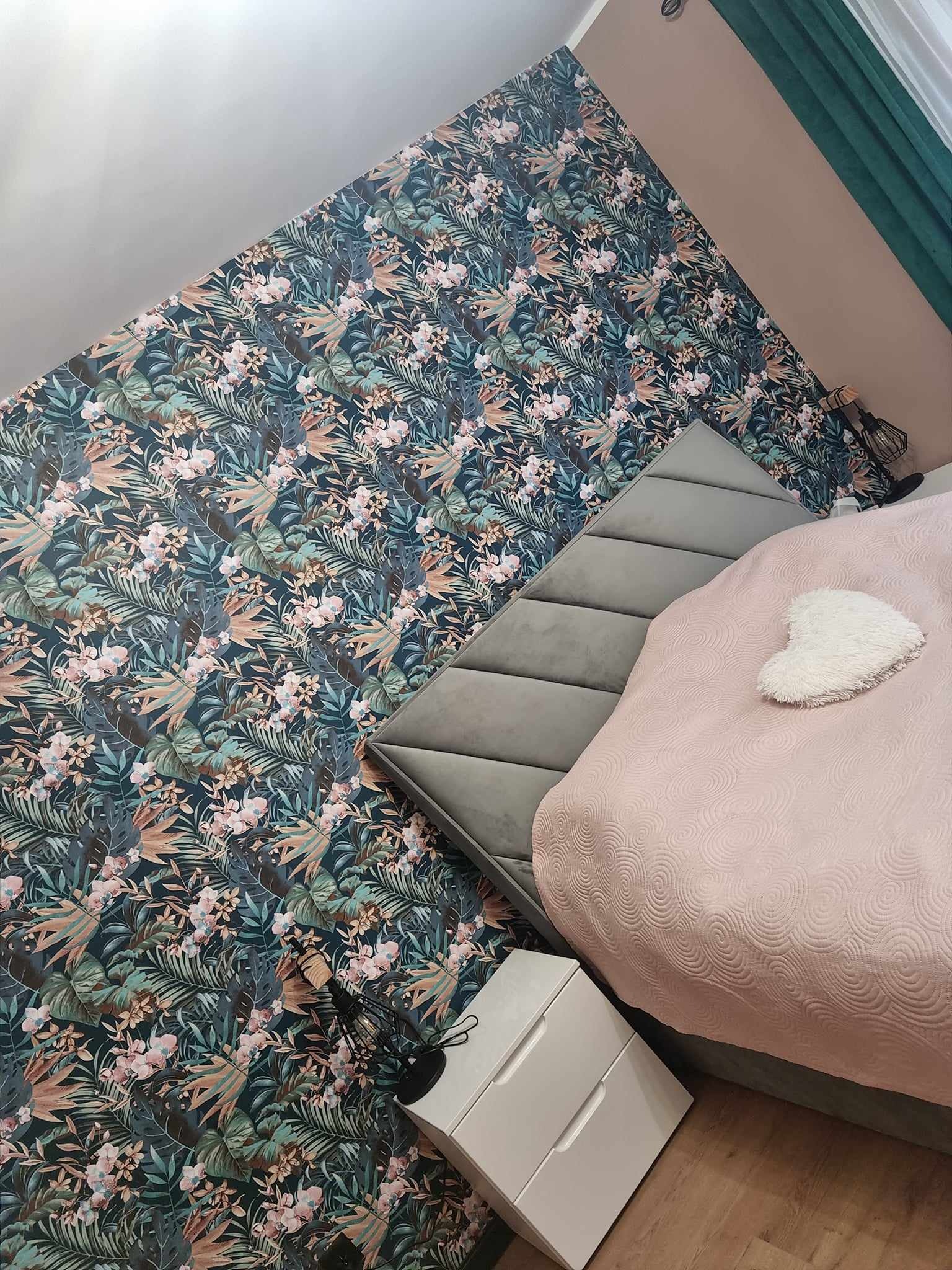 Sypialnia z tapetą w tropikalne wzory, szarą tapicerowaną ścianą za łóżkiem i białą szafką nocną z dwiema szufladami.