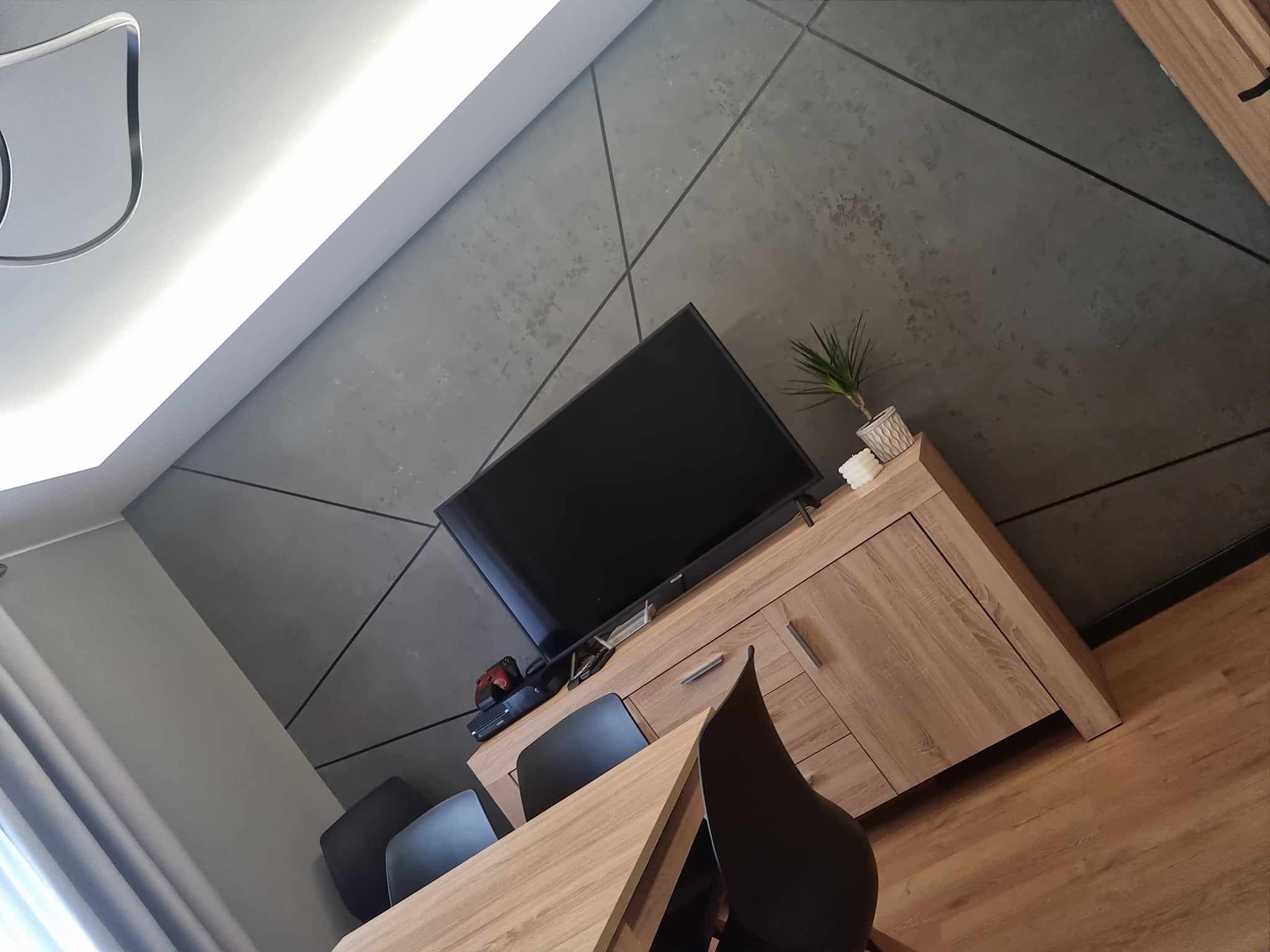 Pokój dzienny z szarą ścianą dekoracyjną z geometrycznym wzorem, drewnianą komodą pod telewizorem i drewnianym stołem z czarnymi krzesłami.