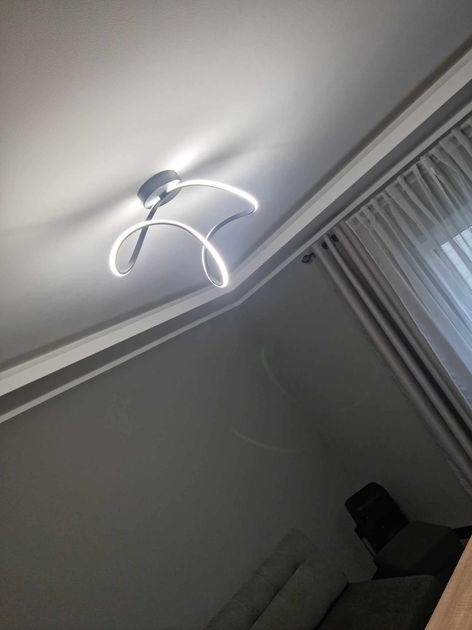 Nowoczesna lampa sufitowa LED o abstrakcyjnym kształcie, świecąca jasnym, białym światłem na tle szarego sufitu i ściany, z widocznymi zasłonami przy oknie.