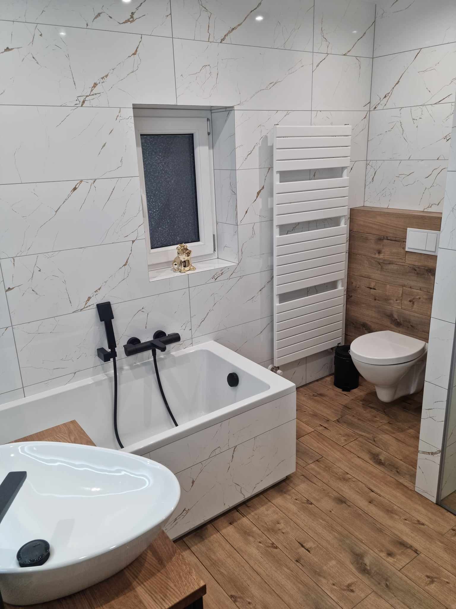Nowoczesna łazienka z białymi płytkami imitującymi marmur, drewnianymi akcentami i czarną armaturą; na parapecie okna stoi złota figurka żaby.