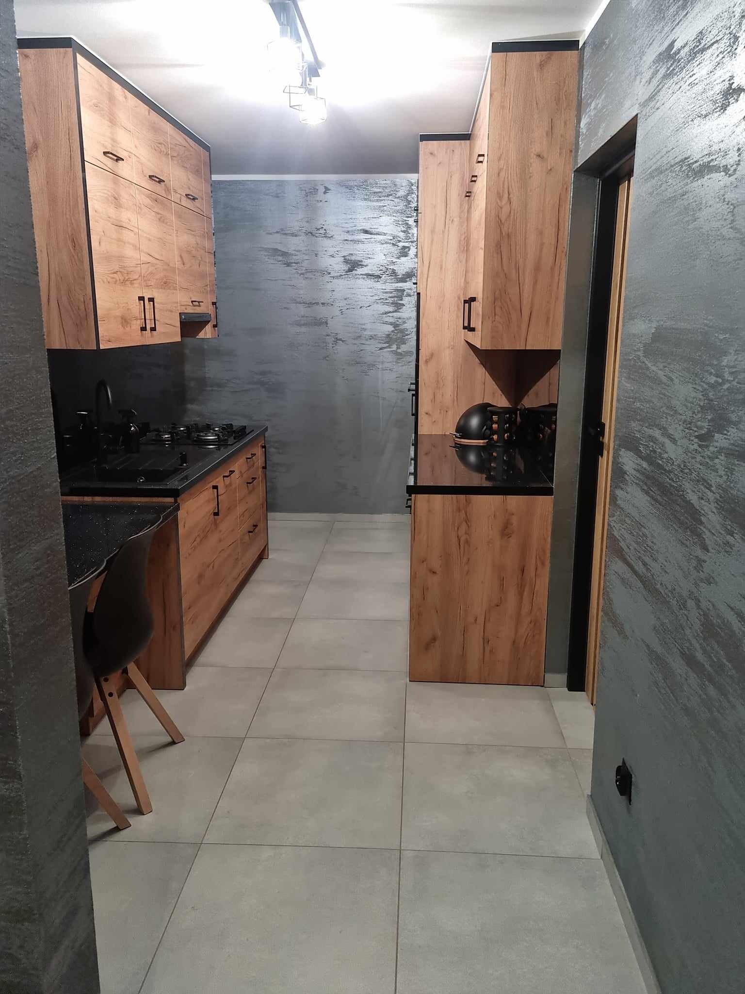Wąska kuchnia z drewnianymi szafkami z czarnymi uchwytami i czarnym blatem, ściany w odcieniach szarości z dekoracyjną fakturą, jasne płytki podłogowe, widok z perspektywy wejścia.