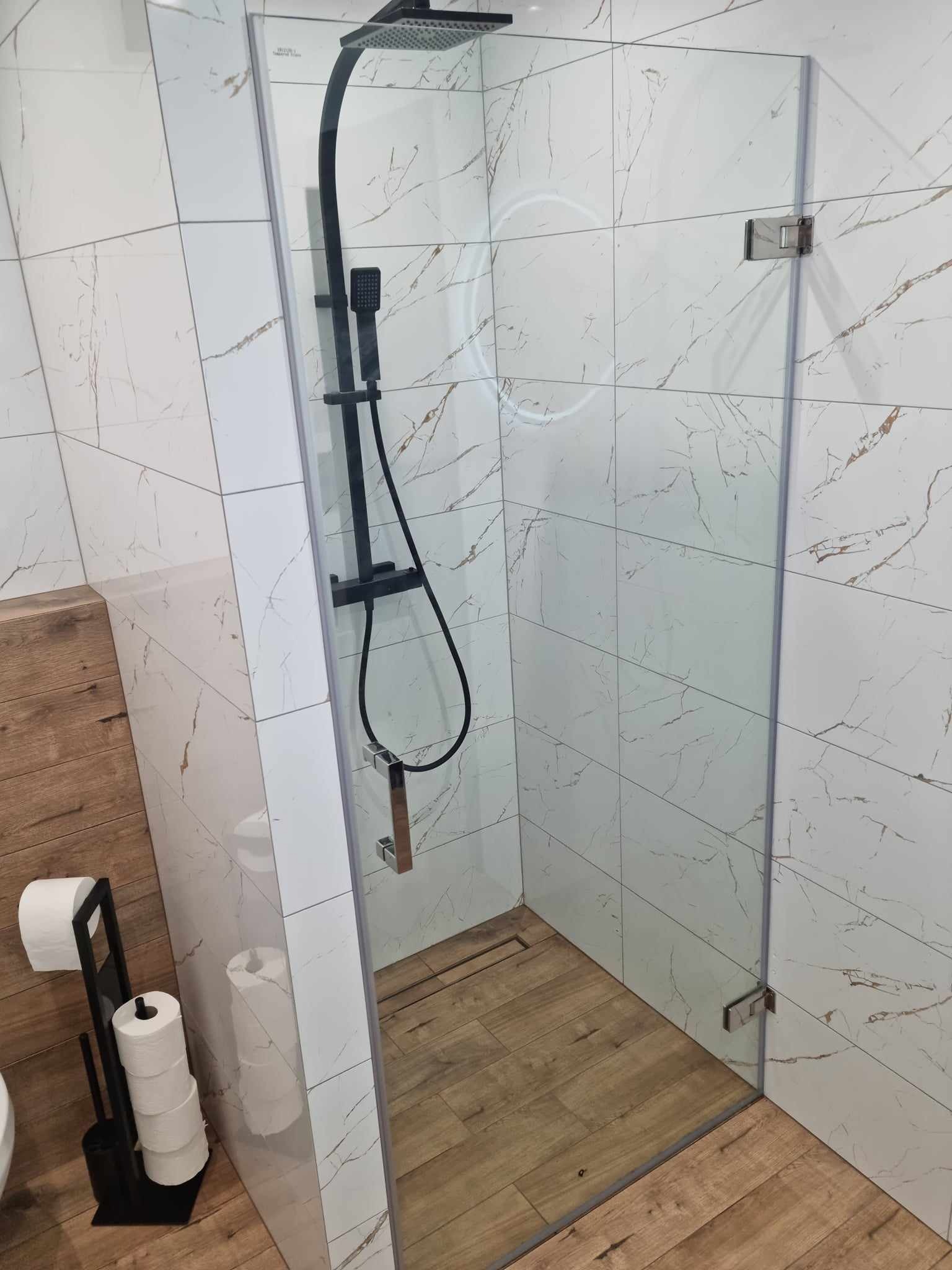 Nowoczesna kabina prysznicowa z czarną armaturą na tle białych płytek imitujących marmur i drewnopodobnej podłogi, widoczny stojak na papier toaletowy.
