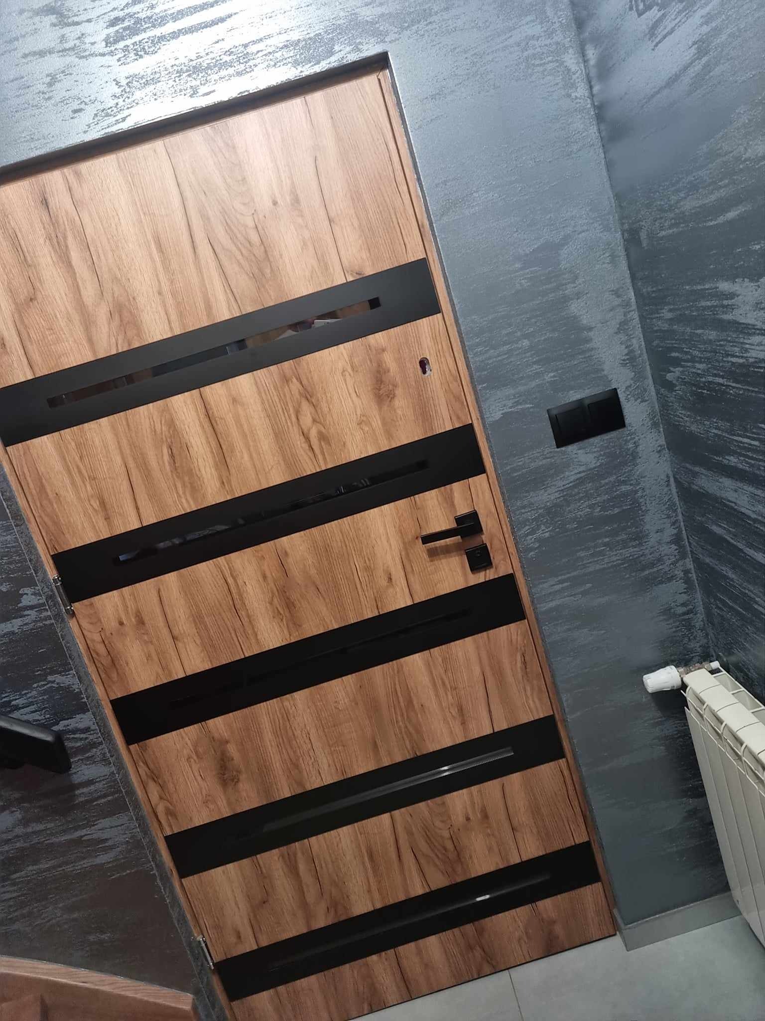 Drewniane drzwi wewnętrzne z czarnymi, poziomymi wstawkami na tle ściany z dekoracyjnym tynkiem w odcieniach szarości, widoczny fragment grzejnika i włącznika światła.