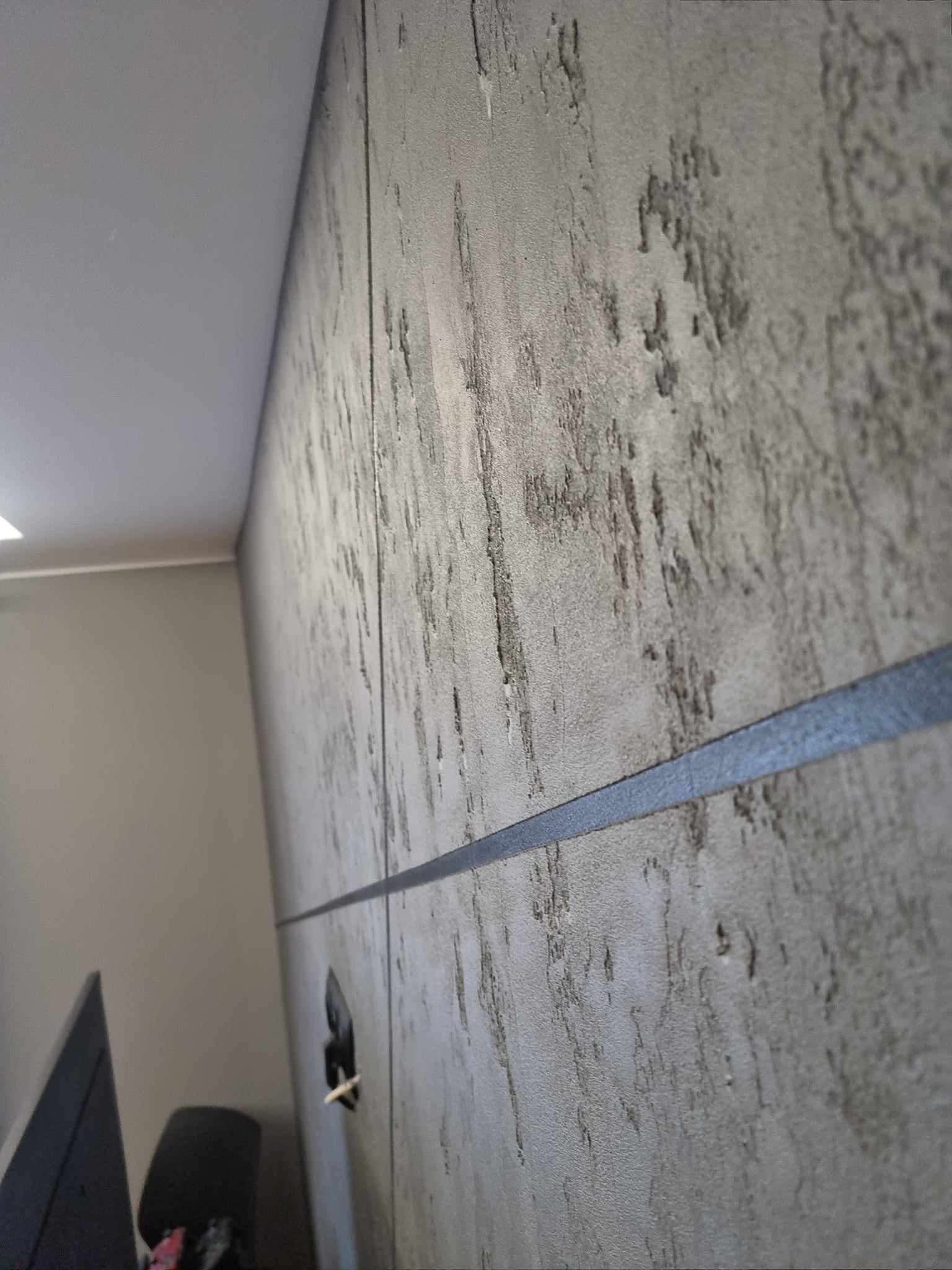 Dekoracyjna szara ściana z efektem betonu i poziomym czarnym pasem, widoczna z bliska pod kątem, z fragmentem sufitu i mebli w tle.