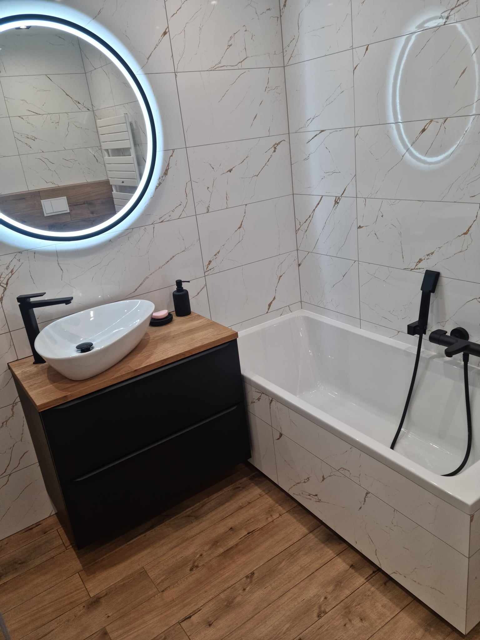 Nowoczesna łazienka z owalną umywalką nablatową na czarnej szafce z drewnianym blatem, białą wanną obudowaną płytkami imitującymi marmur, okrągłym lustrem z podświetleniem LED i czarną armaturą.