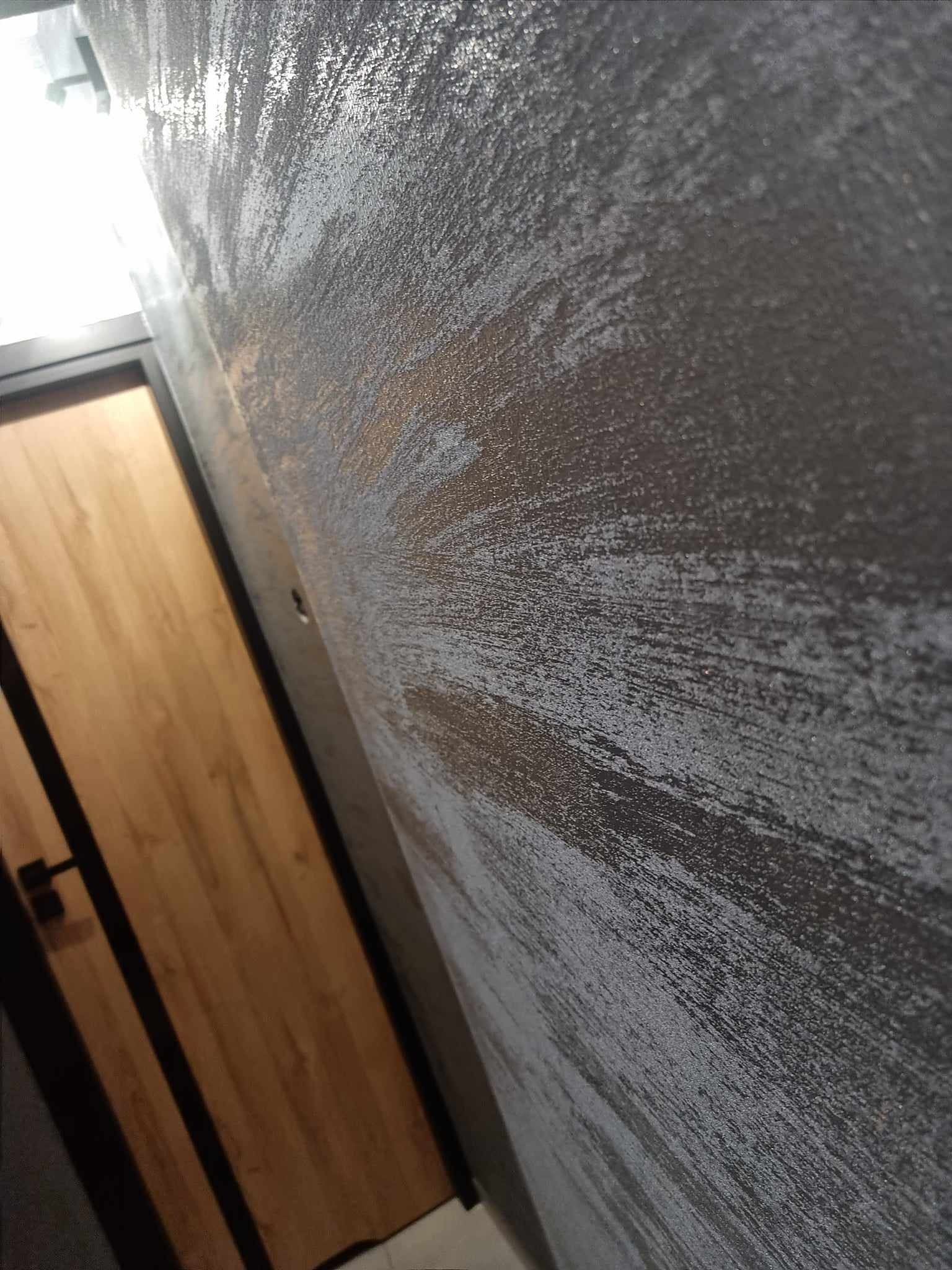 Ściana z dekoracyjną szarą strukturą, obok widoczna drewniana ościeżnica drzwiowa z czarnymi elementami wykończeniowymi, ujęcie z dołu.