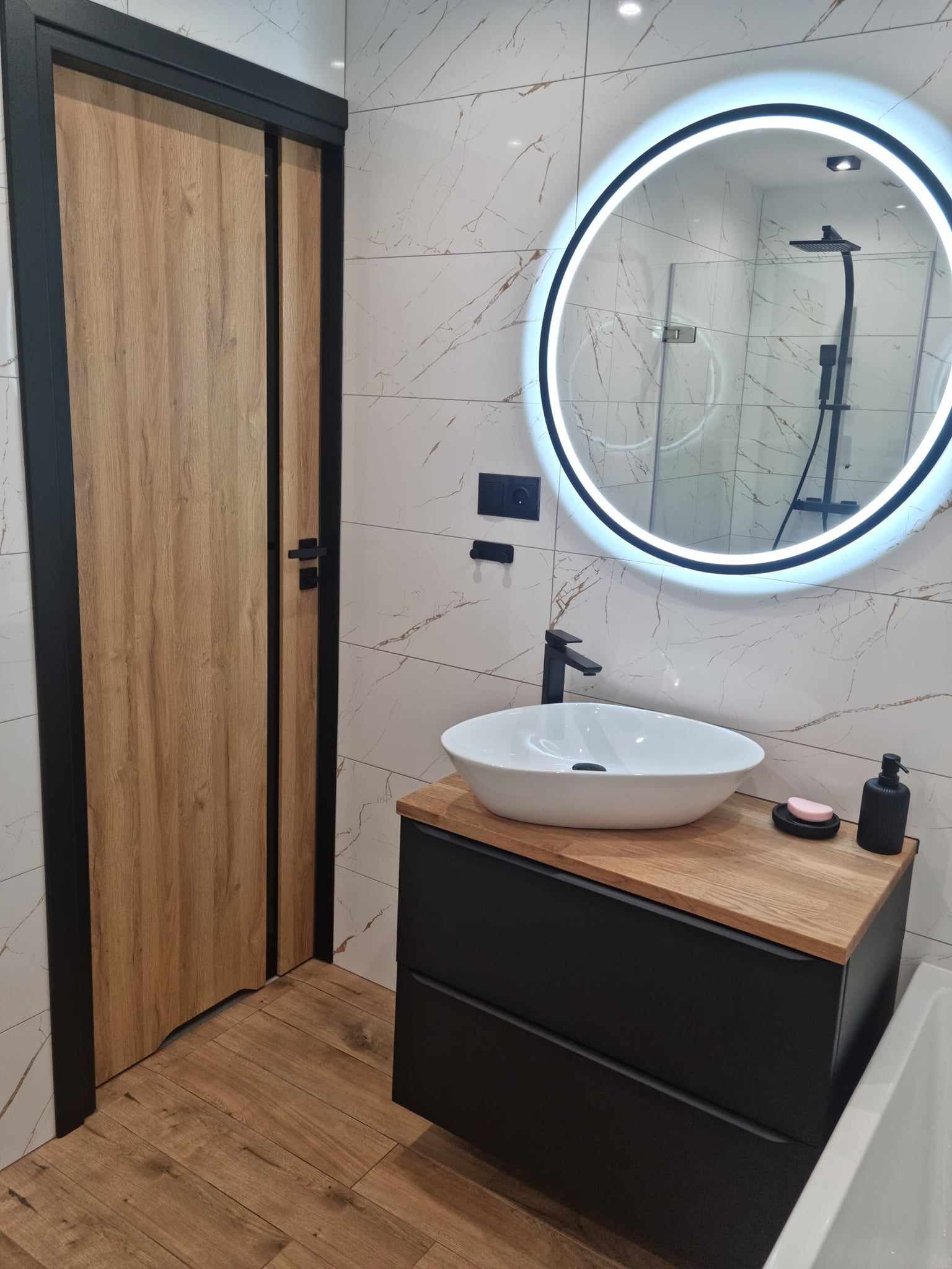 Nowoczesna łazienka w Dobrzycy z drewnianą podłogą i drzwiami, marmurowe płytki ścienne, okrągłe lustro z podświetleniem LED, umywalka nablatowa na czarnej szafce z drewnianym blatem i widoczną...