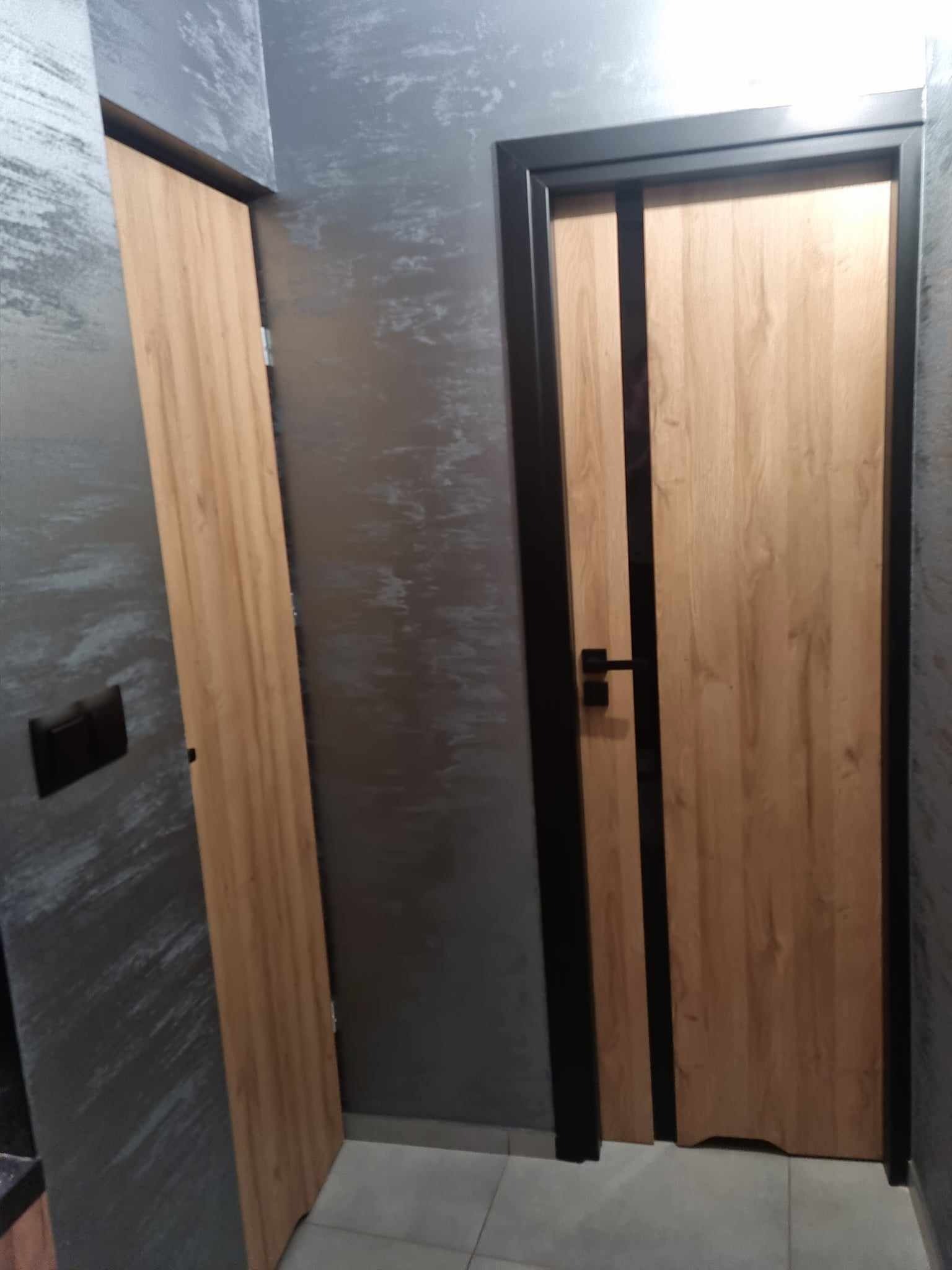 Dwie drewniane drzwi wewnętrzne z czarnymi ościeżnicami i klamkami na tle szarej ściany z efektem dekoracyjnym, widoczne płytki podłogowe.