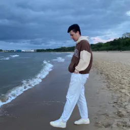 Młody mężczyzna w brązowej bluzie z kapturem i białych spodniach spaceruje po piaszczystej plaży wzdłuż brzegu morza, patrząc w d&oacute;ł na fale. W tle widoczne budynki przemysłowe i zachmurzone niebo.