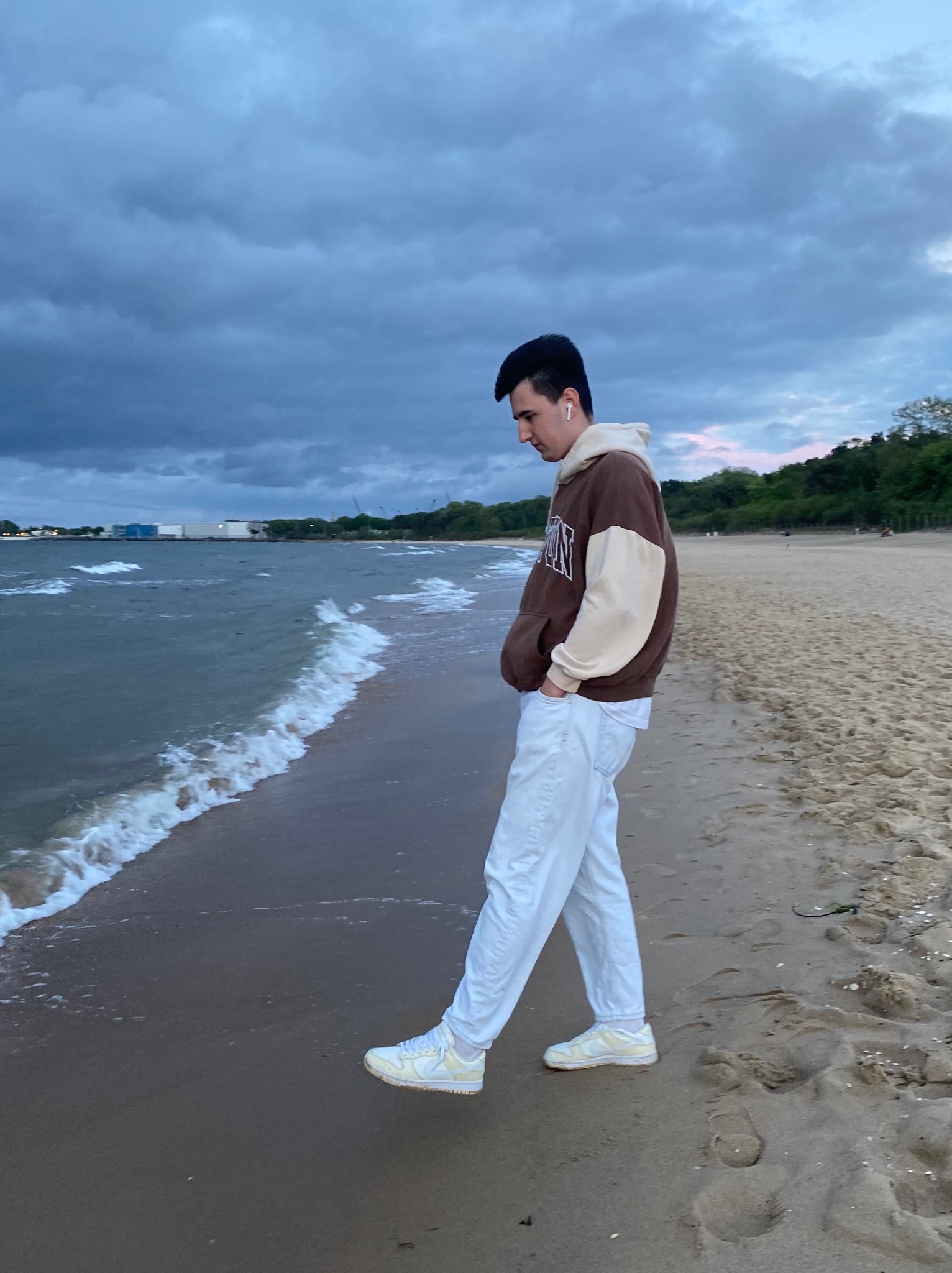 Młody mężczyzna w brązowej bluzie z kapturem i białych spodniach spaceruje po piaszczystej plaży wzdłuż brzegu morza, patrząc w dół na fale. W tle widoczne budynki przemysłowe i zachmurzone niebo.