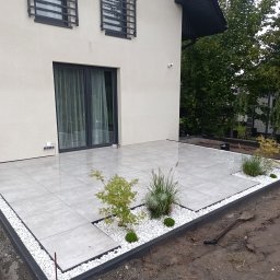 SEB-POL Sebastian Chmielarz - Nowoczesny taras z jasnoszarej kostki, obrzeża z białego kamienia i czarnych krawężników. W tle dom z czarnymi ramami okiennymi i zielenią. Widoczna łopata.