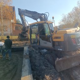 SEB-POL Sebastian Chmielarz - Żółto-szara koparka Volvo EW140D przy wykopie z betonowymi ścianami, w tle dom i drzewa. Widoczni pracownicy. Słoneczny dzień.