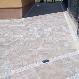 SEB-POL Sebastian Chmielarz - Ułożona kostka brukowa w odcieniach szarości z ozdobnymi obrzeżami z drobnych kamieni. W tle pracownicy podczas układania kostki. Widok z góry.