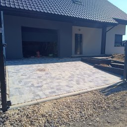 SEB-POL Sebastian Chmielarz - Ułożona kostka brukowa na podjeździe do garażu nowoczesnego domu z szarą elewacją. Widoczny fragment ogrodzenia i paleta z kostką. Jasne, słoneczne oświetlenie.
