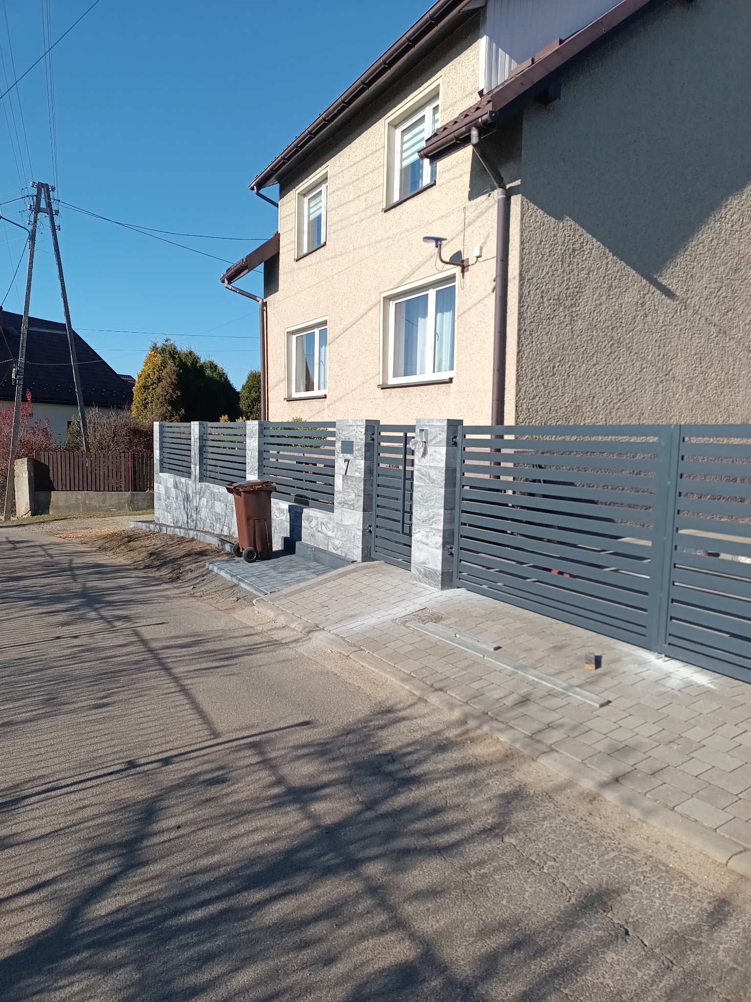 Nowoczesne ogrodzenie panelowe w kolorze grafitowym z bramą, wykończone płytkami imitującymi kamień, przed domem z beżową elewacją w słoneczny dzień.