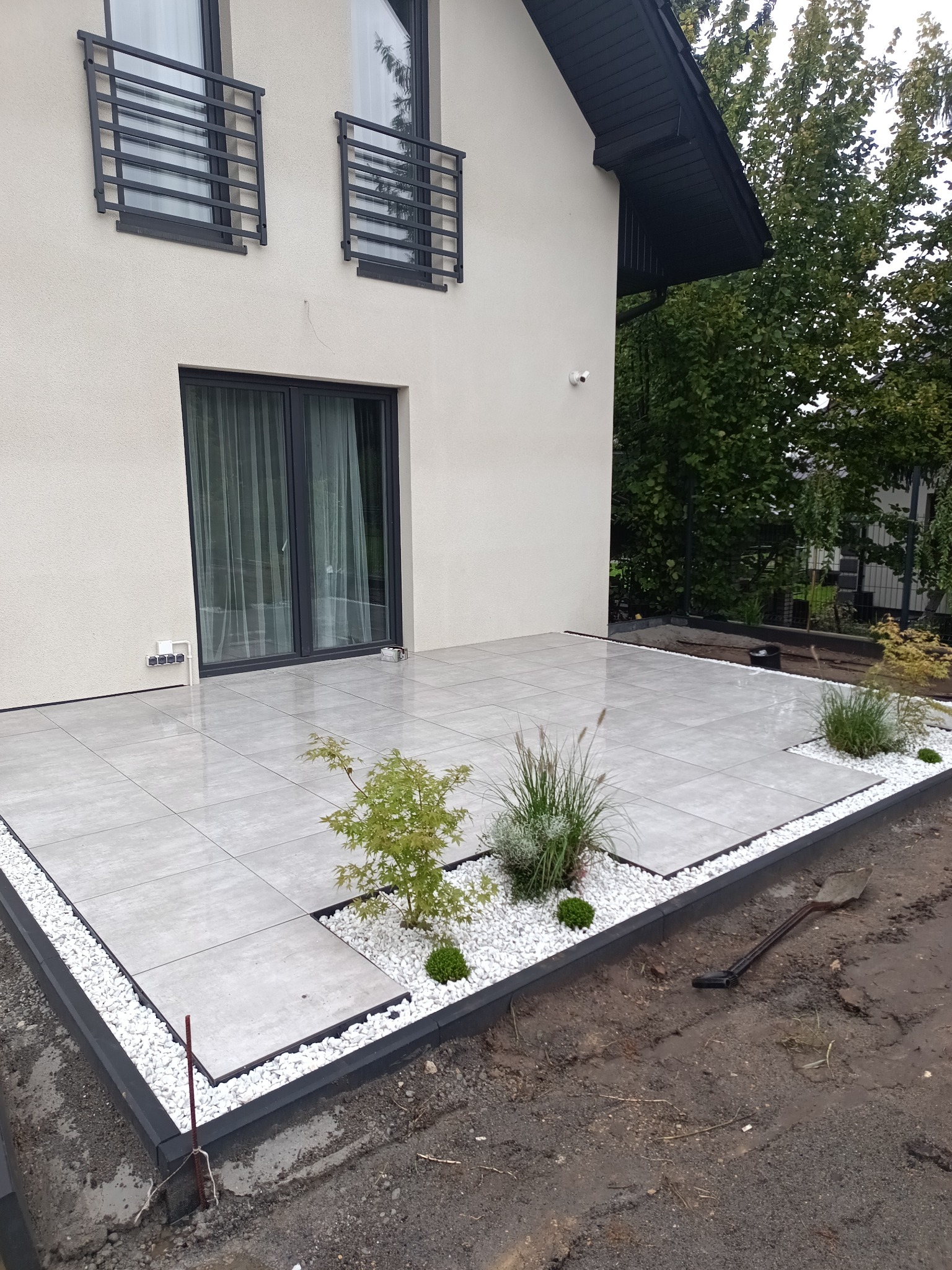 Nowoczesny taras z jasnoszarej kostki, obrzeża z białego kamienia i czarnych krawężników. W tle dom z czarnymi ramami okiennymi i zielenią. Widoczna łopata.