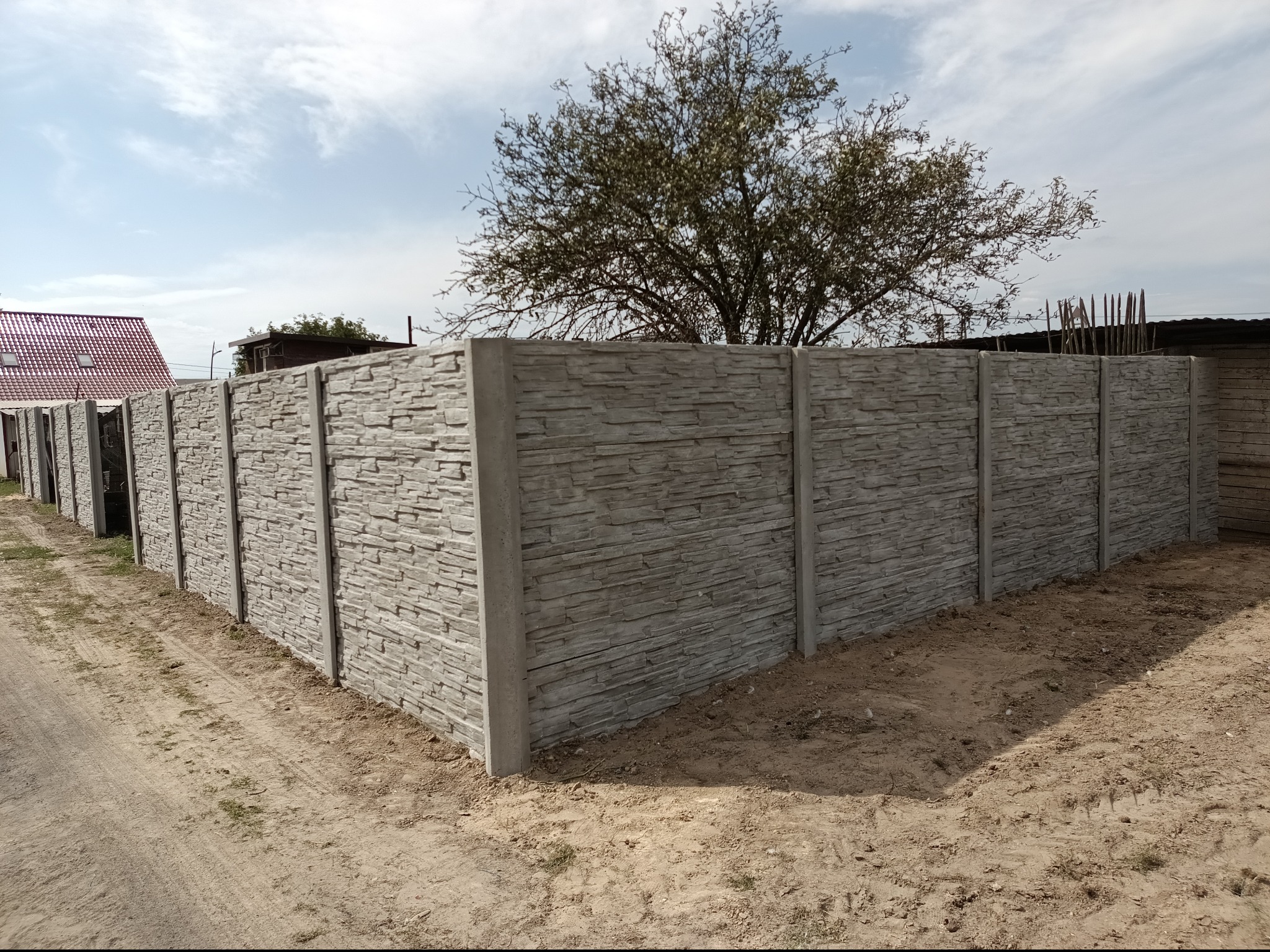 Długi, szary, betonowy płot z fakturą kamienia, oddzielający posesję. Widoczne słupki i panele. W tle drzewo i fragment dachu. Ujęcie z poziomu gruntu.