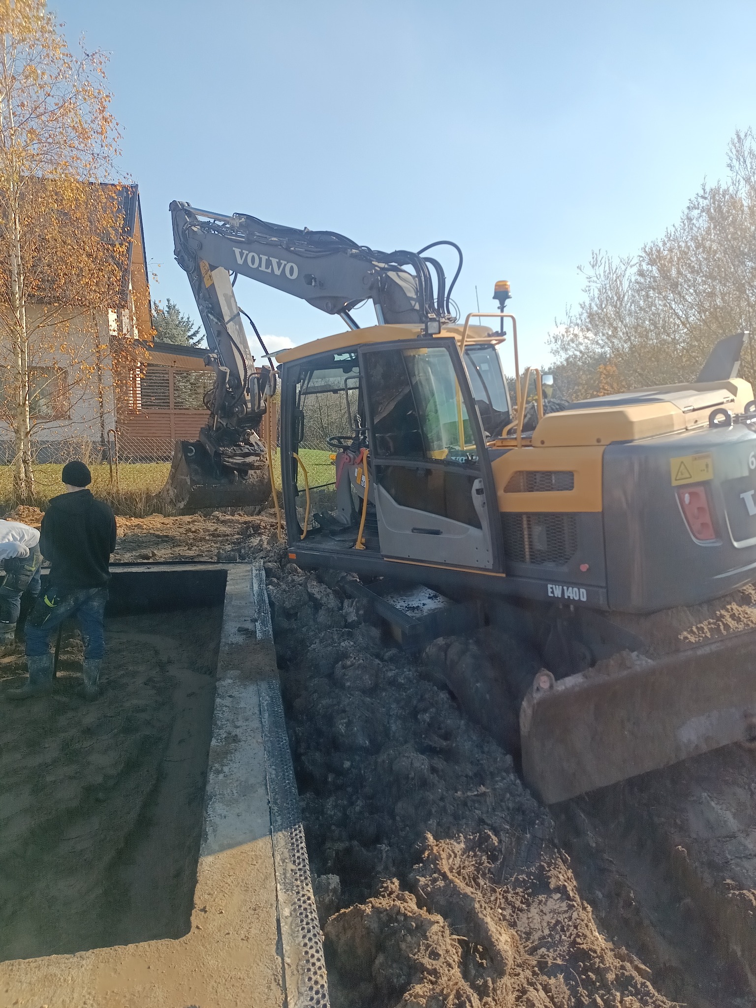 Żółto-szara koparka Volvo EW140D przy wykopie z betonowymi ścianami, w tle dom i drzewa. Widoczni pracownicy. Słoneczny dzień.