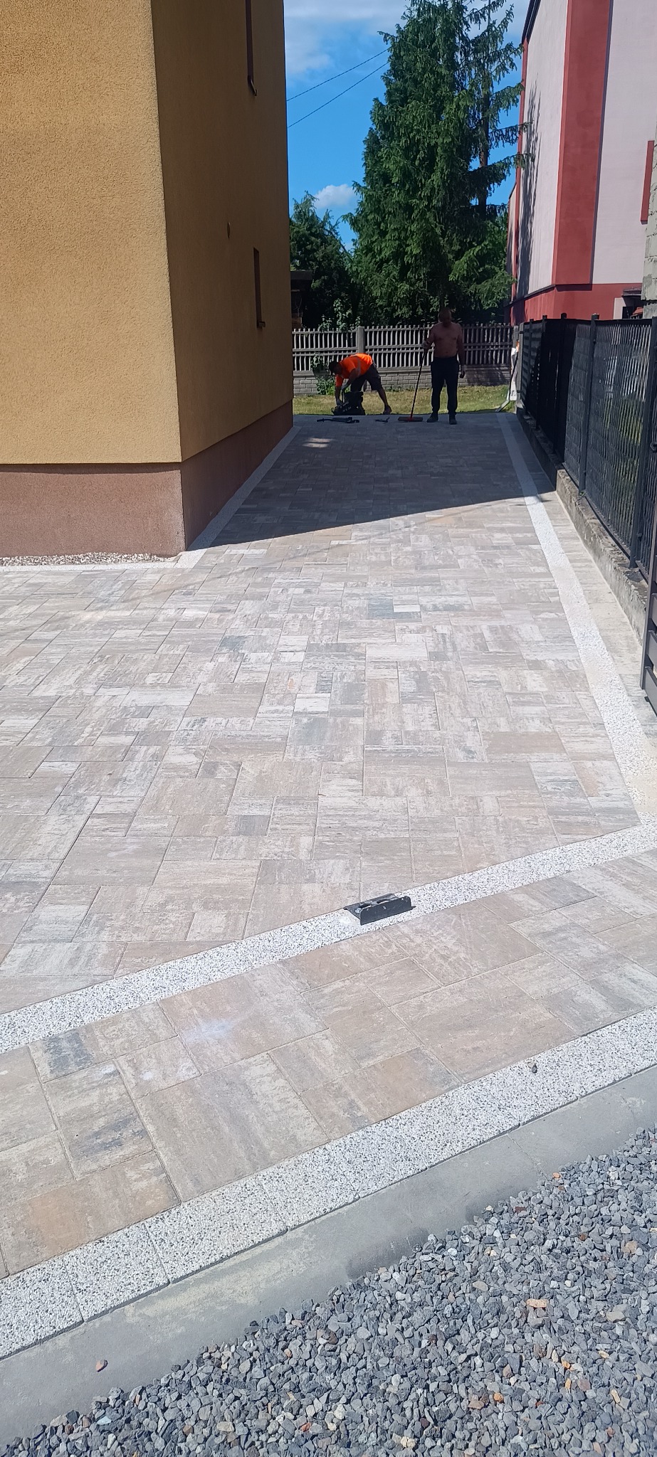 Ułożona kostka brukowa w odcieniach szarości z ozdobnymi obrzeżami z drobnych kamieni. W tle pracownicy podczas układania kostki. Widok z góry.