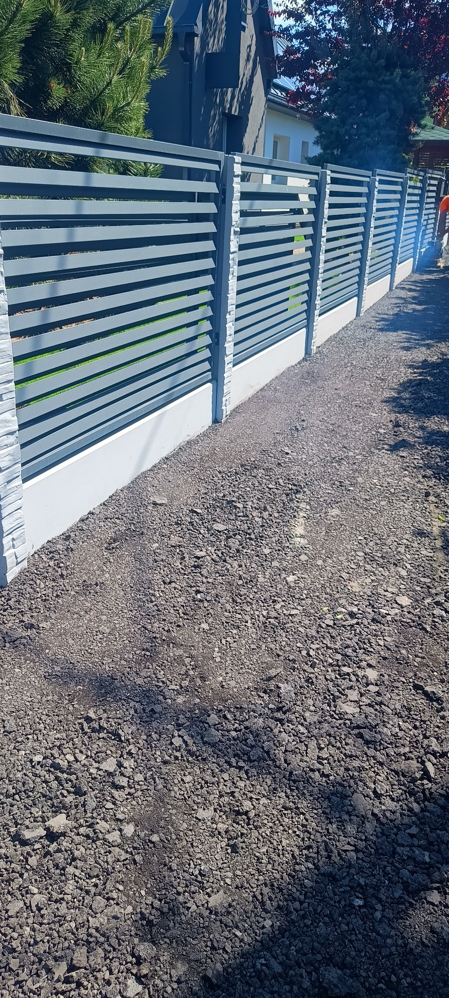 Nowoczesne ogrodzenie panelowe w kolorze szarym z podmurówką, widok z perspektywy gruntu. Słupki wykończone imitacją kamienia. Tło: dom jednorodzinny i zieleń.