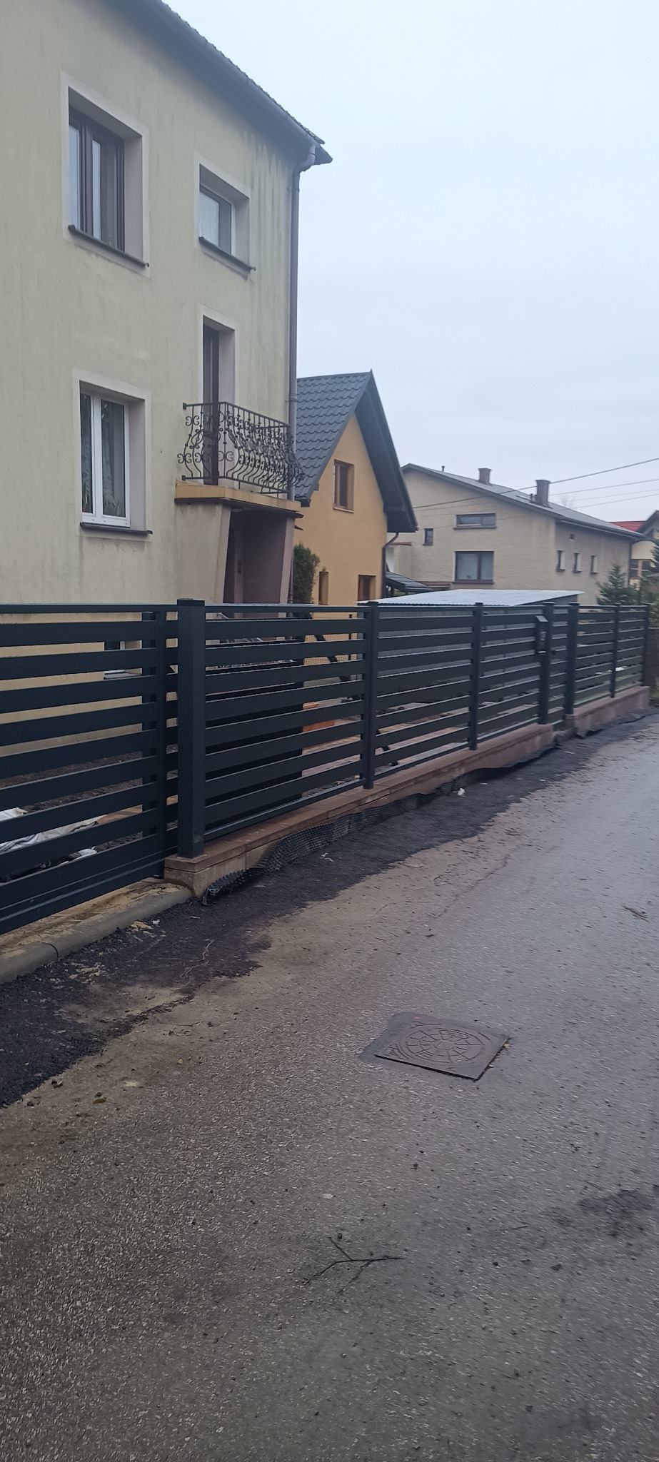 Nowoczesne, poziome ogrodzenie panelowe w kolorze antracytowym, zamontowane przed domem. Widoczne elementy montażowe i podmurówka. Ujęcie z poziomu ulicy.