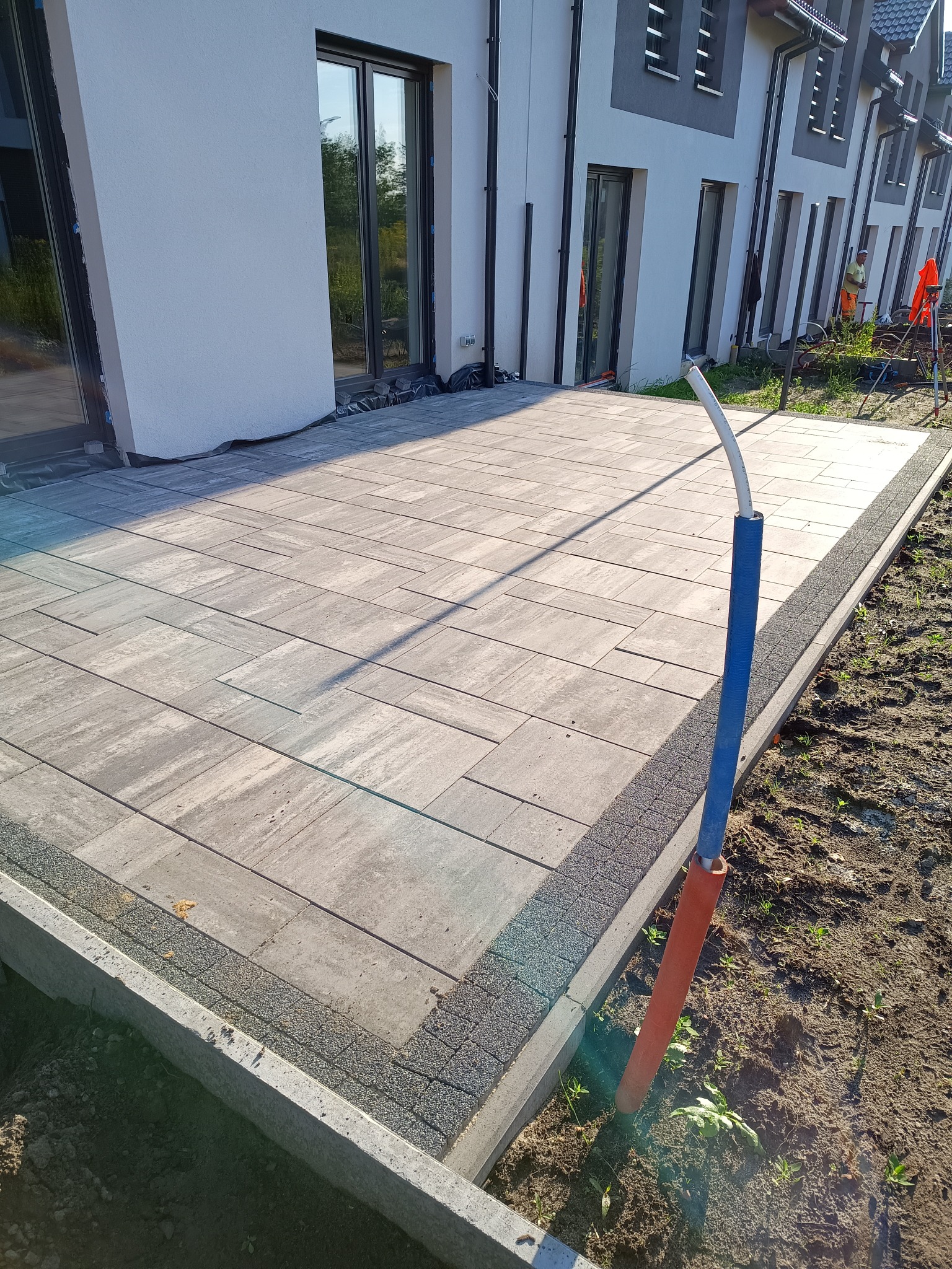 Ułożona kostka brukowa na tarasie domu jednorodzinnego, widoczne obrzeża i fragment instalacji. Jasne, naturalne światło.