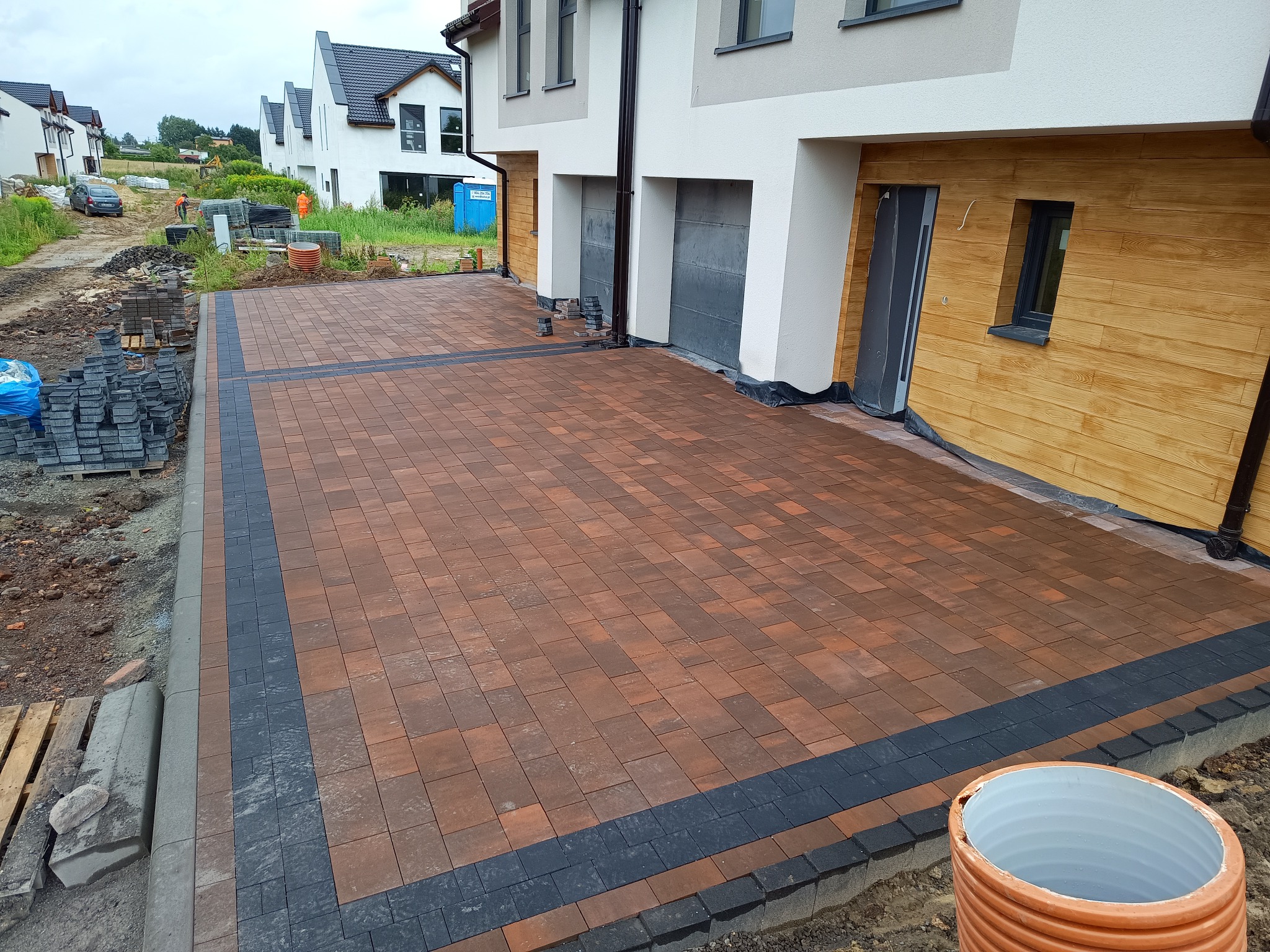 Ukończony podjazd z kostki brukowej w odcieniach brązu i grafitu, z obrzeżami. W tle domy jednorodzinne i materiały budowlane.