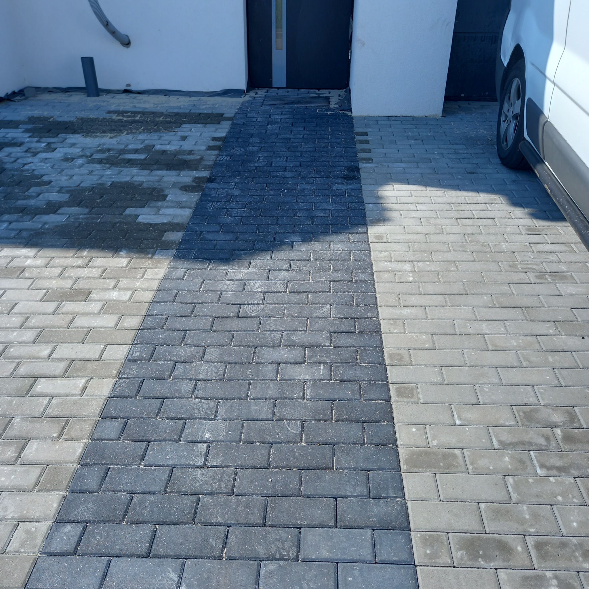 Ułożona kostka brukowa w dwóch odcieniach szarości tworząca wzór. Widoczny fragment elewacji budynku i koło samochodu. Kostka ułożona równo i estetycznie.