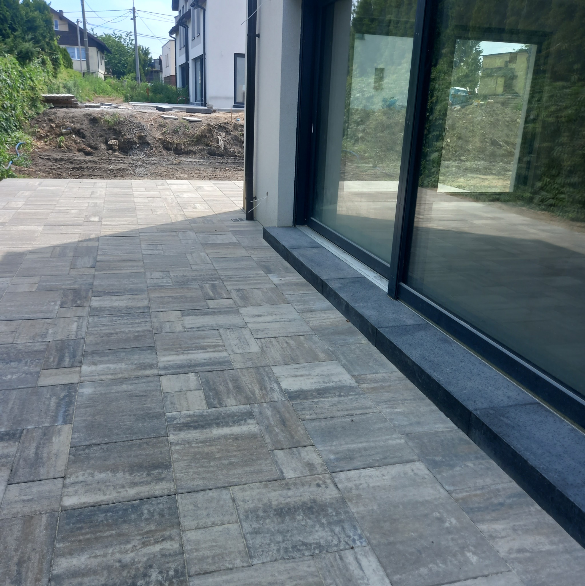 Ułożona kostka brukowa w odcieniach szarości, przylegająca do nowoczesnego budynku z dużymi oknami. W tle widoczny teren z ziemią i roślinnością.