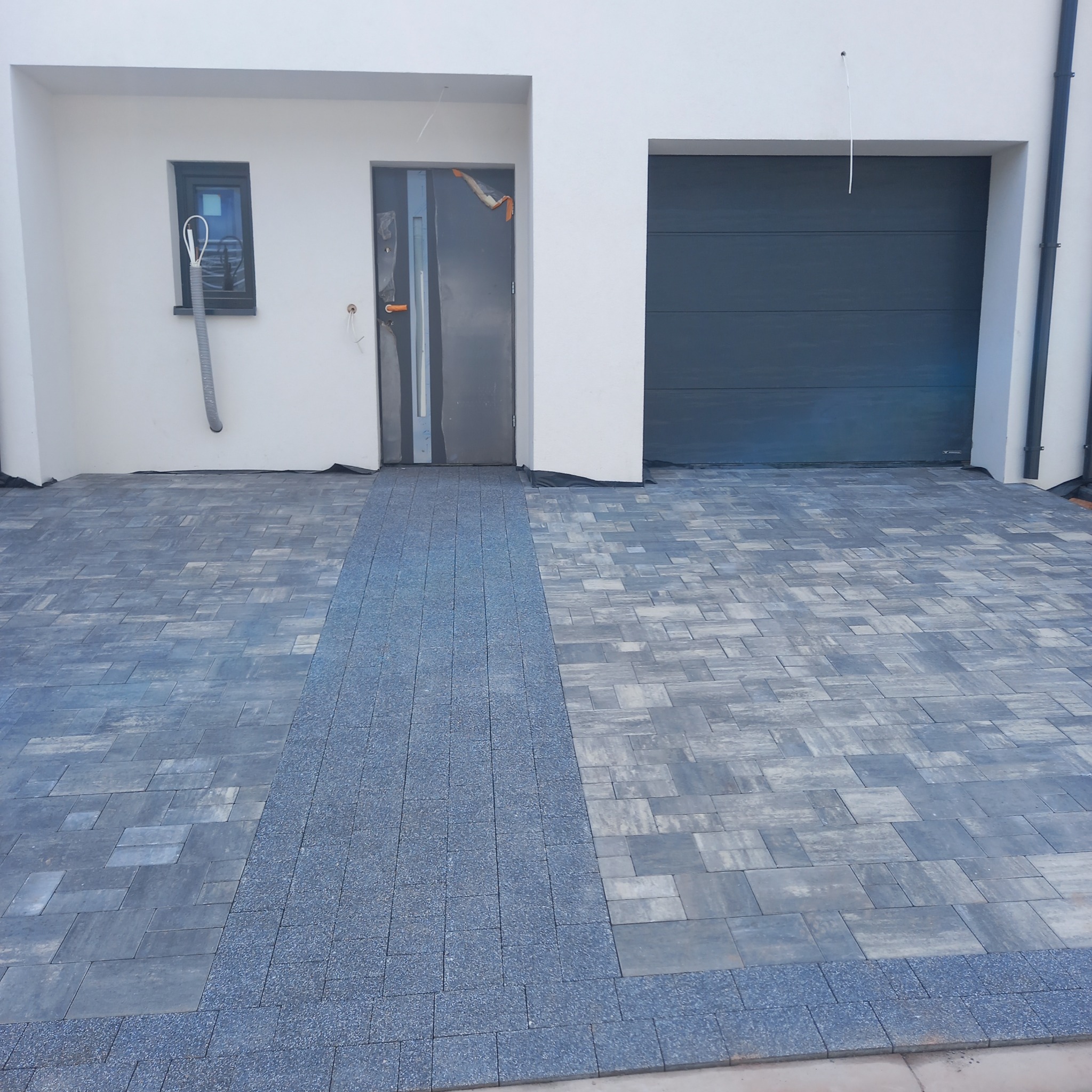 Ułożona, szara kostka brukowa przed nowoczesnym domem z garażem i drzwiami wejściowymi. Widoczny wzór ułożenia kostki i jej faktura.