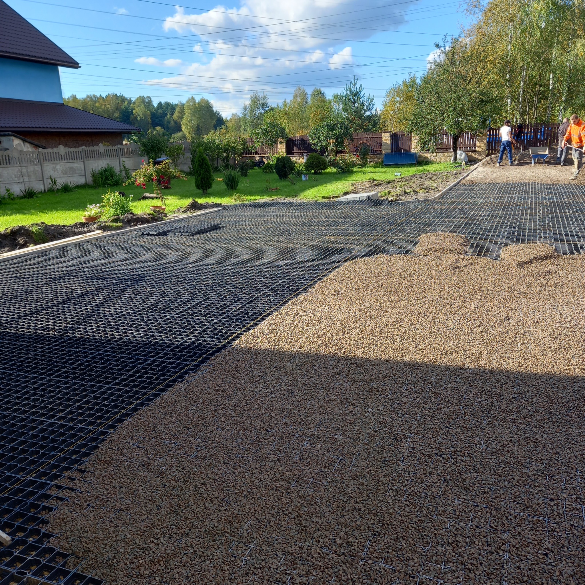 Przygotowanie podłoża pod nawierzchnię w Jaworzno: czarna siatka i częściowo wysypany żwir. W tle pracownicy i zieleń ogrodu.