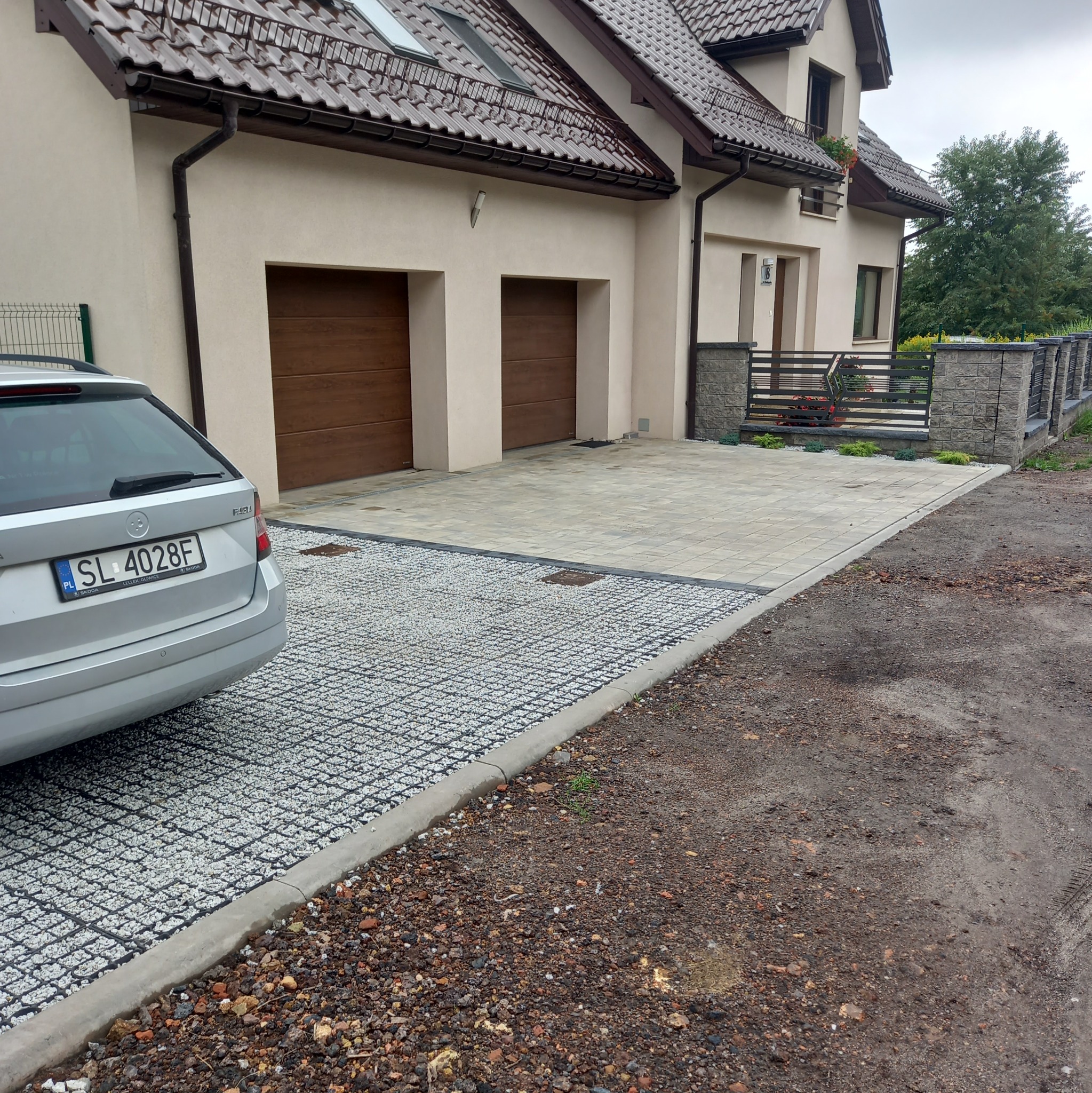 Nowoczesny podjazd z kostki brukowej i ażurowych płyt wypełnionych grysem, prowadzący do garażu w beżowym domu z brązowymi bramami. Częściowo widoczny samochód.