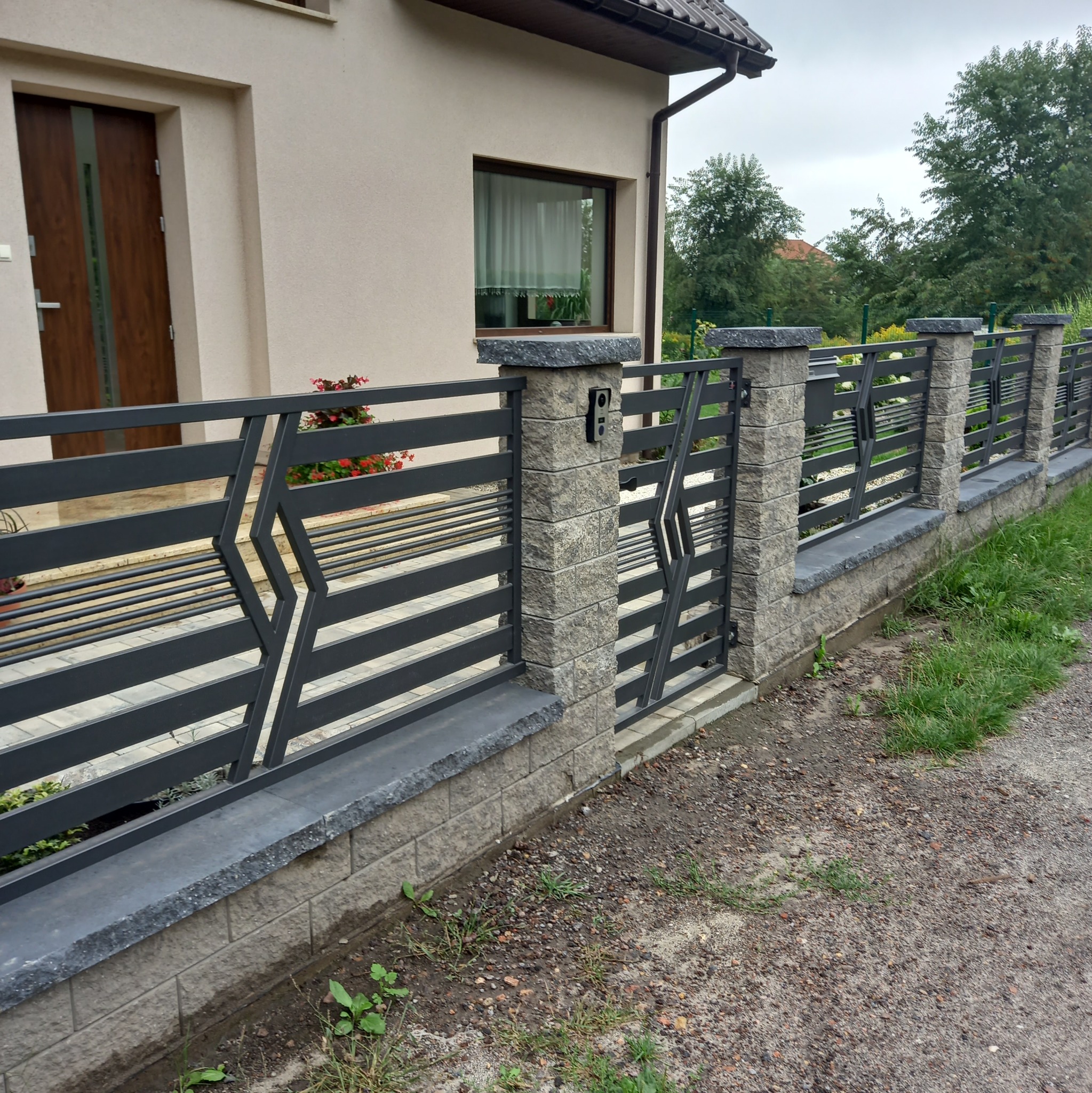 Nowoczesne ogrodzenie panelowe z bramką, z elementami z szarego kamienia i metalu, na tle domu z beżową elewacją i brązowymi drzwiami.