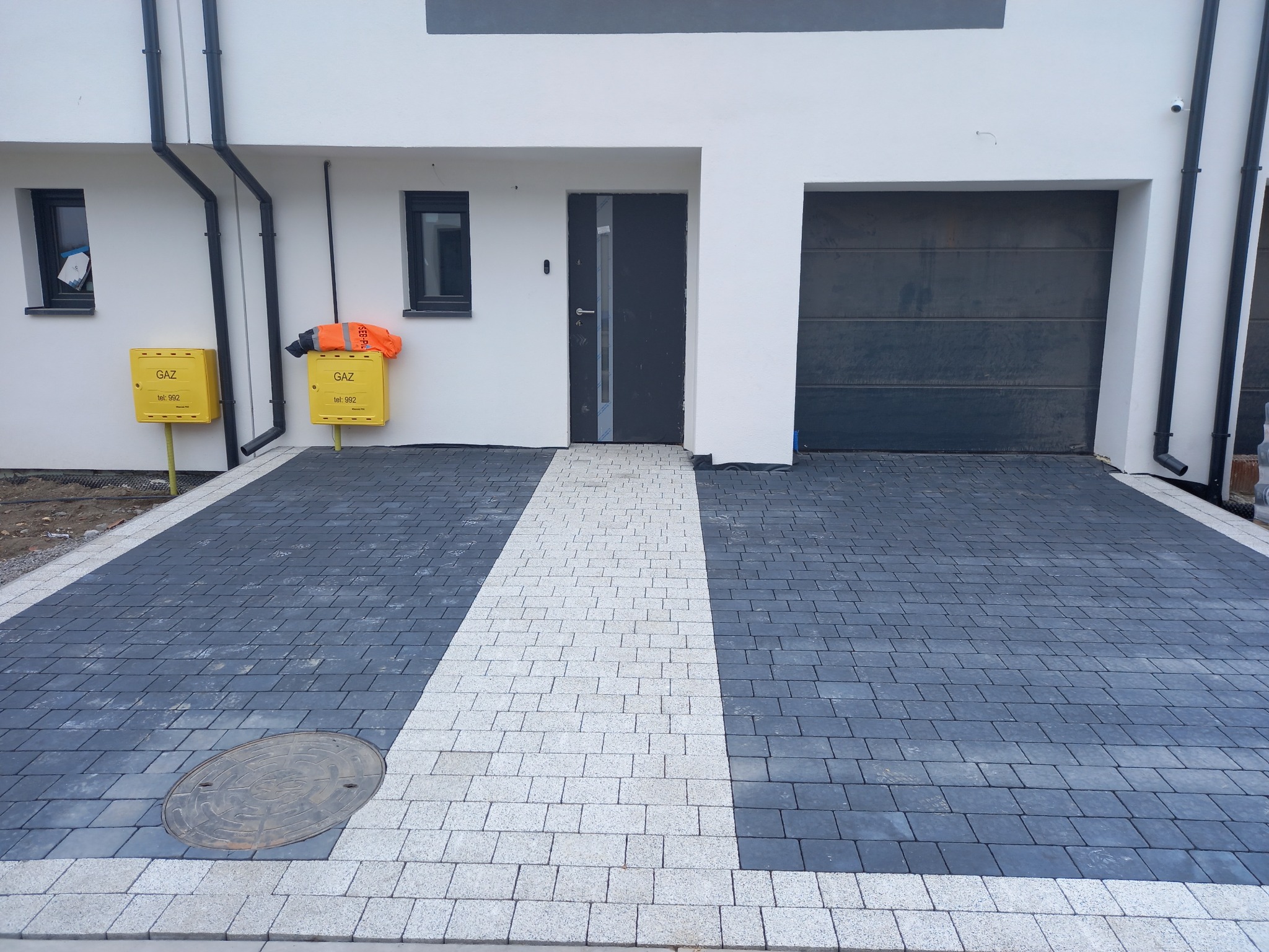 Nowoczesny podjazd z kostki brukowej w odcieniach szarości i bieli, prowadzący do domu z garażem. Widoczne studzienki kanalizacyjne i skrzynki gazowe.