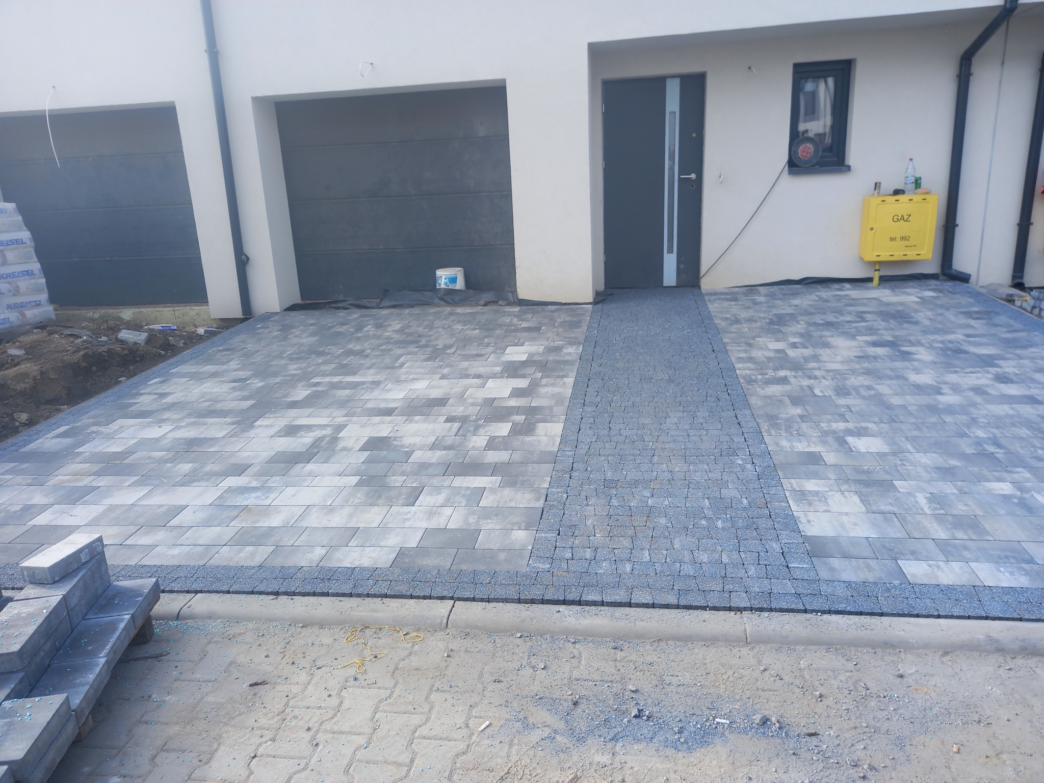 Nowoczesny podjazd z szarej kostki brukowej ułożonej wzorem prostokątnym, z pasem drobnej kostki prowadzącym do drzwi wejściowych. Widoczny fragment domu z garażem.