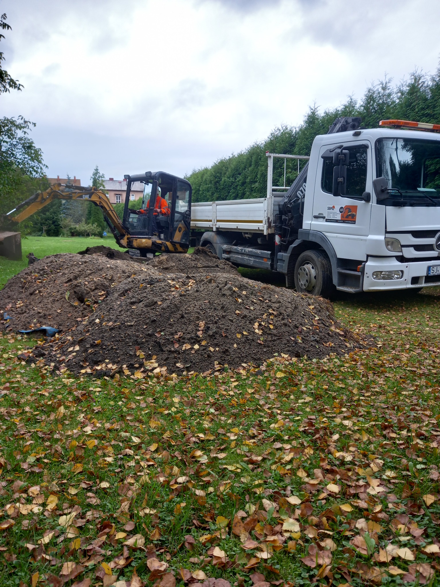 Żółta minikoparka z operatorem w pomarańczowej kamizelce obok białej ciężarówki z dźwigiem, na trawniku z liśćmi, podczas prac ziemnych. Pochmurne niebo.