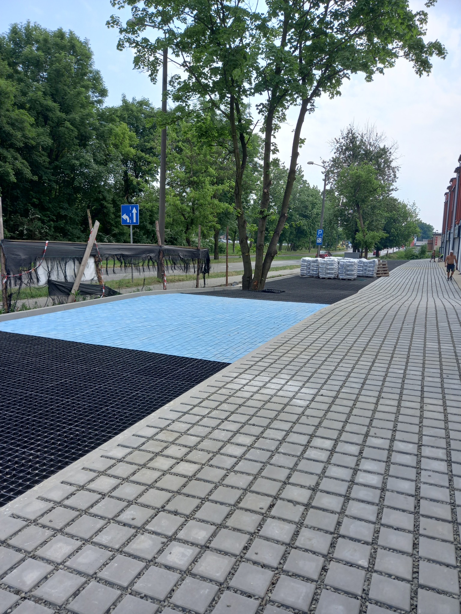 Nowa nawierzchnia z kostki brukowej w szarym odcieniu, z fragmentami w kolorze niebieskim i czarnym, otoczona zielenią drzew. Widoczne worki z materiałem budowlanym.