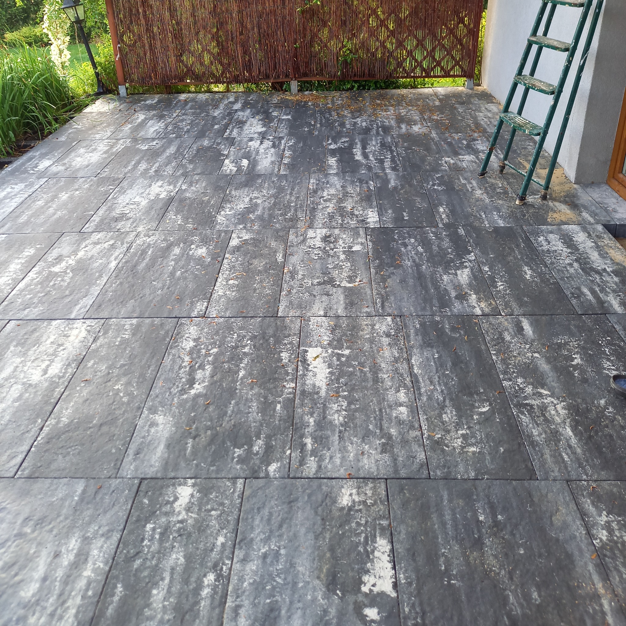 Ułożona, szara kostka brukowa na tarasie z bambusową osłoną i zieloną drabiną opartą o ścianę. Widok z góry, lekko z ukosa.