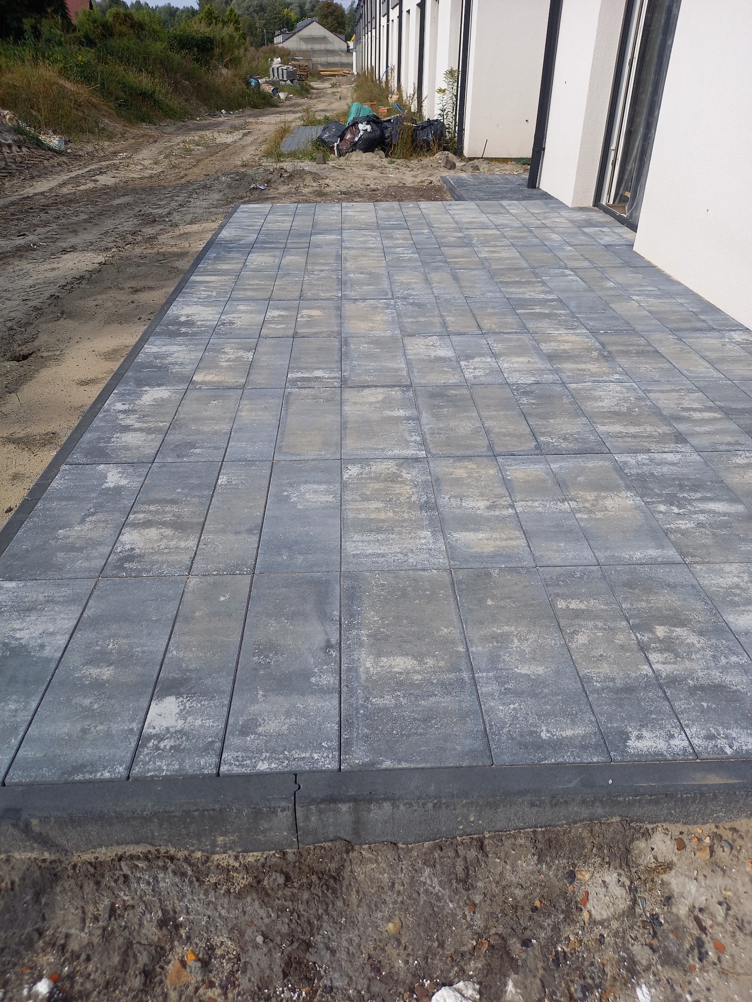 Ułożona, szara kostka brukowa na posesji, widok z góry. Krawężnik oddziela kostkę od ziemi. W tle budynek z białymi ścianami i okna.