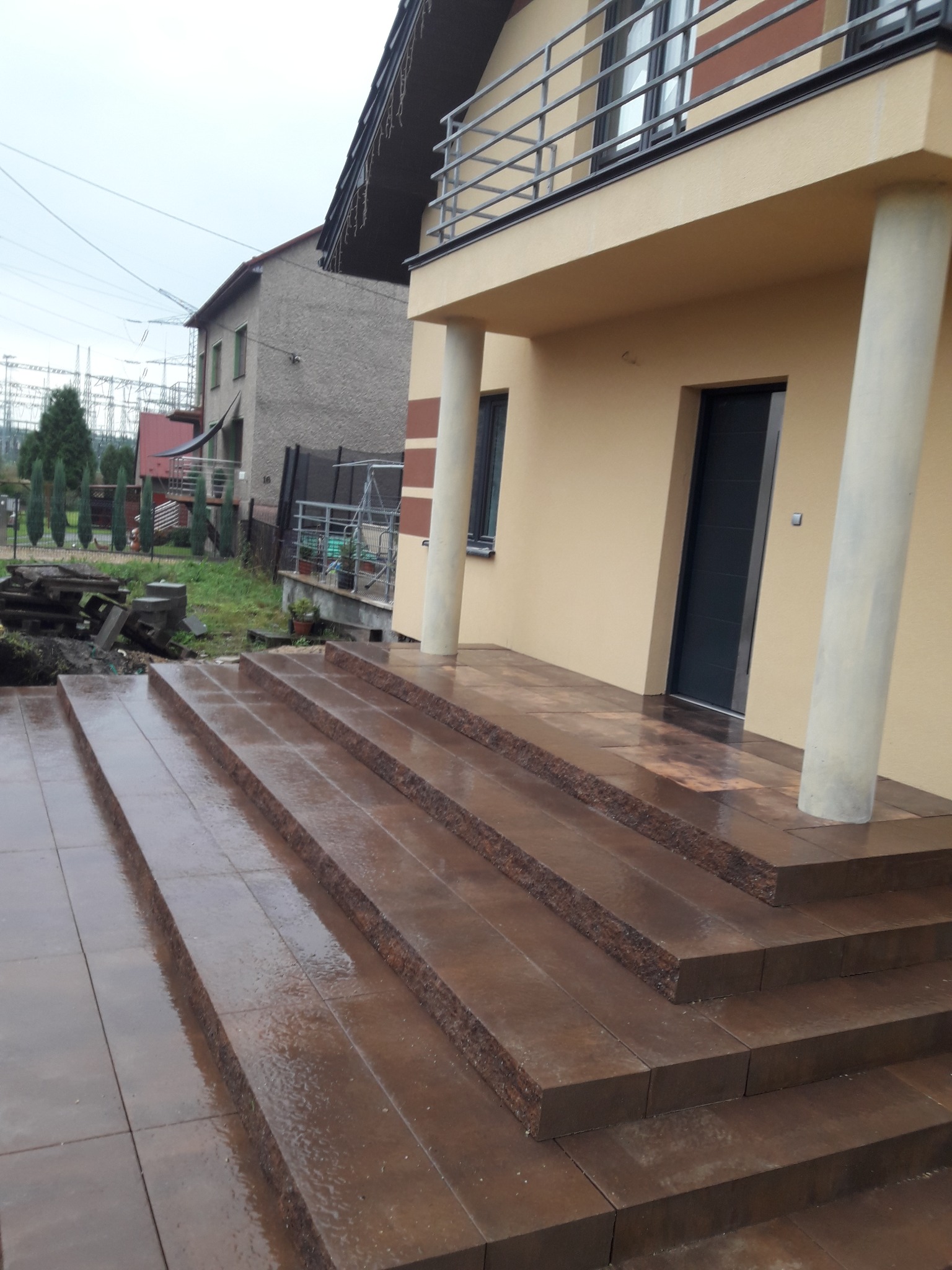 Ułożone schody z kostki brukowej prowadzące do domu z balkonem. Widoczne detale wykończenia i sąsiedni budynek w tle. Kostka mokra po deszczu.