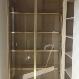 Wysoka, przeszklona szafa wnękowa z złotymi ramami i szklanymi półkami, widoczna drabina wewnątrz, białe ściany i wykładzina.