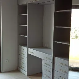 Szara zabudowa meblowa z szafami, p&oacute;łkami i biurkiem, złote uchwyty, podłoga zabezpieczona kartonem.