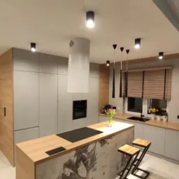 Nowoczesna kuchnia z szarymi frontami, drewnianym blatem i wyspą z betonowym wykończeniem i motywem kwiatowym, oświetlona wiszącymi lampami i punktowymi reflektorami.