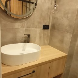 Firma remontowo-wykończeniowa - Nowoczesna łazienka z okrągłym lustrem z podświetleniem, wiszącą szafką z drewnianym blatem, ceramiczną umywalką nablatową i minimalistyczną armaturą w kolorze czarnym, uzupełniona szklaną lampą...