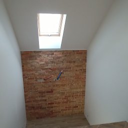 Firma remontowo-wykończeniowa - Klatka schodowa z ceglaną ścianą i drewnopodobnymi płytkami ułożonymi w jodełkę. Widoczne okno dachowe i wystające rury instalacji. Nowoczesne wykończenie.