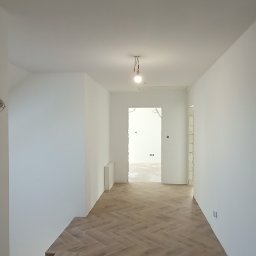 Firma remontowo-wykończeniowa - Korytarz po remoncie z parkietem ułożonym w jodełkę. Ściany pomalowane na biało, widoczne schody i przejście do innego pomieszczenia. W tle surowa żarówka.