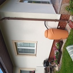 Firma remontowo-wykończeniowa - Elewacja domu po malowaniu z brązową rynną i zbiornikiem na deszczówkę. Widoczne okna z kwiatami i kamienny chodnik. Jasna kolorystyka, spokojne otoczenie.